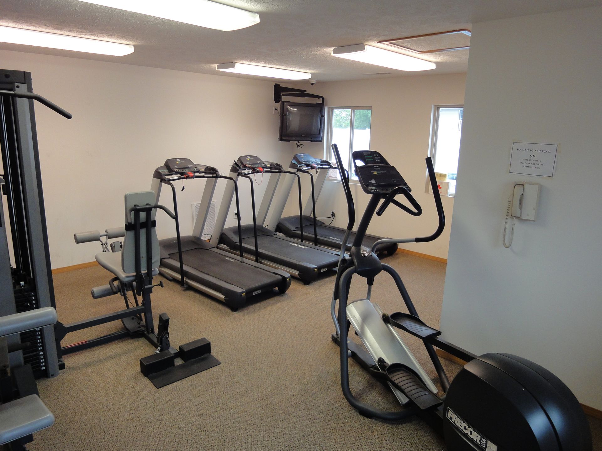Gym with treadmills, elliptical, and weight machine. Beige carpet, neutral walls, TV on wall.