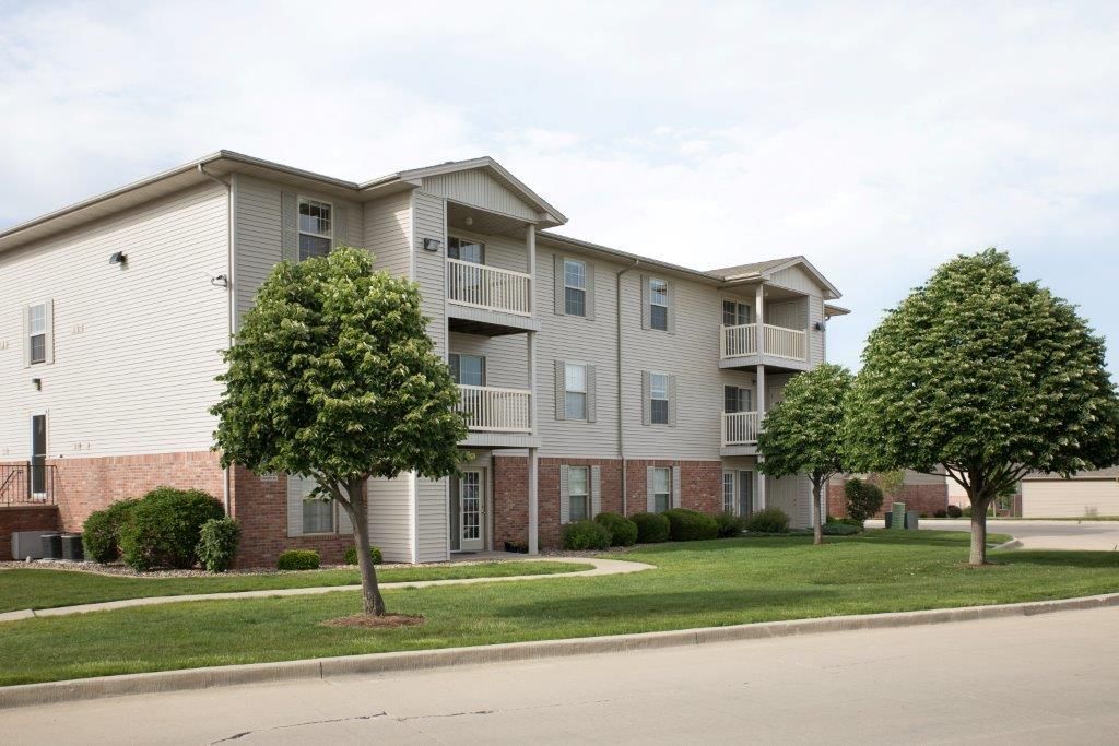 Apartment building with beige siding, red brick base, balconies, and green lawn.