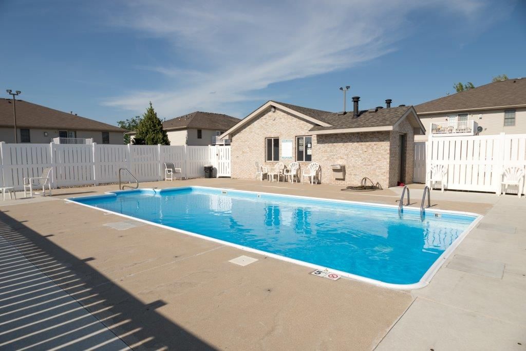 Swimming pool surrounded by a concrete patio and white fence; a brick building in the background under a blue sky.