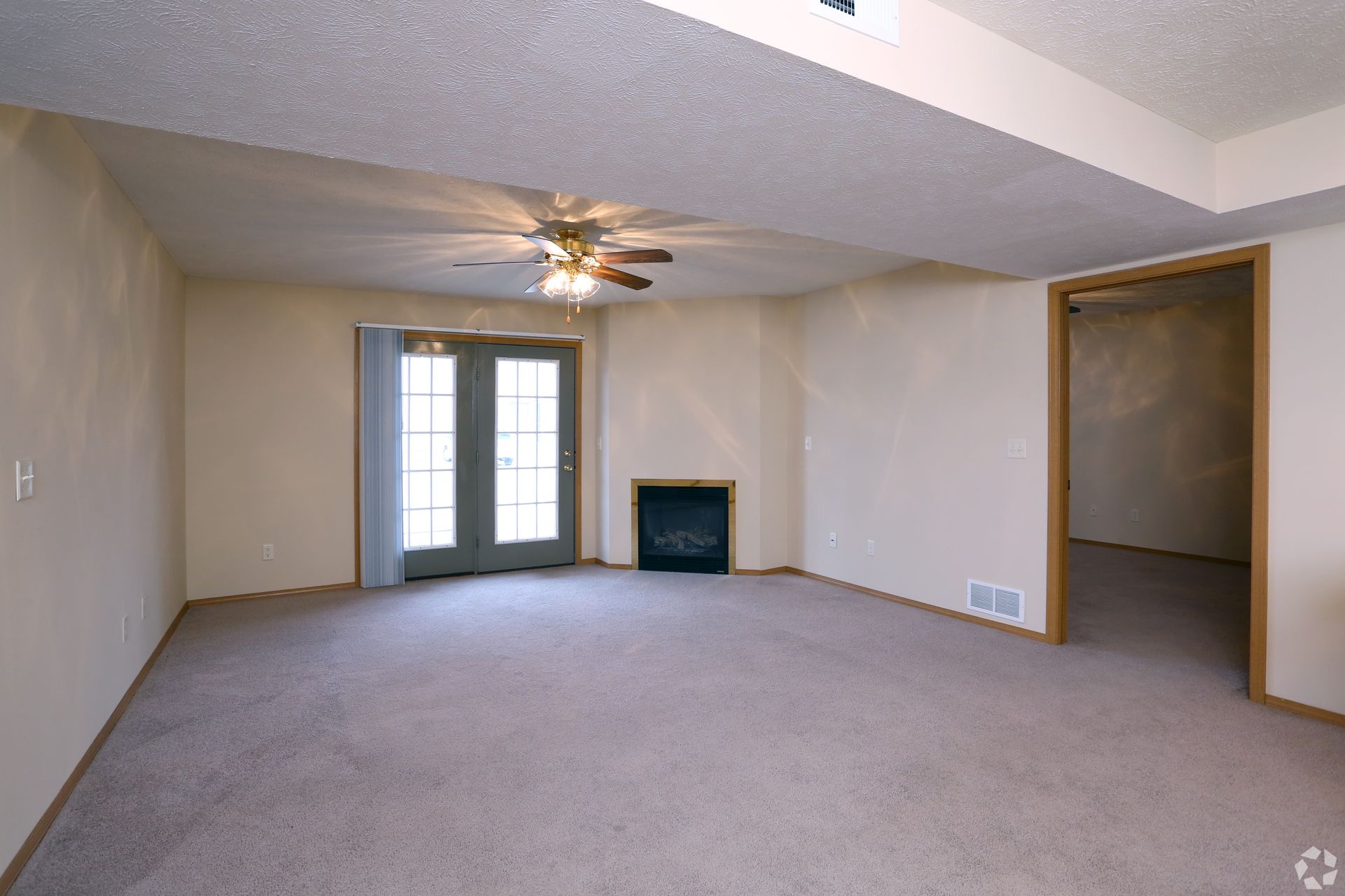 Empty carpeted room with double doors, fireplace, and open doorway; ceiling fan and off-white walls.