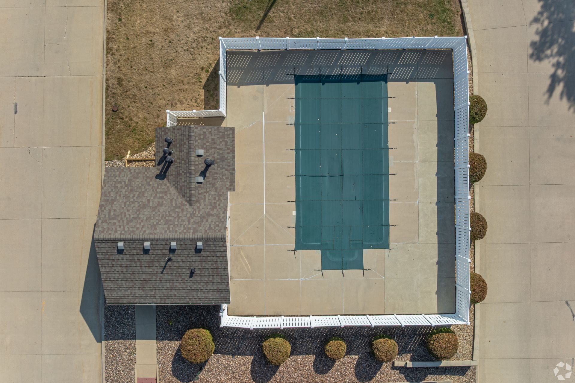 Overhead view of a rectangular pool with a green cover next to a building with a brown roof and surrounding concrete.