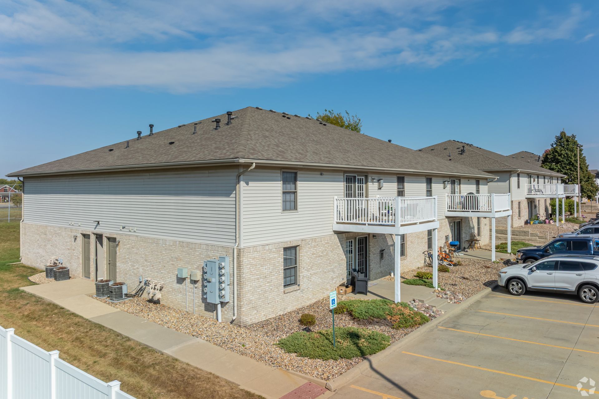 Two-story apartment complex with beige siding, balconies, and parking in a sunny setting.