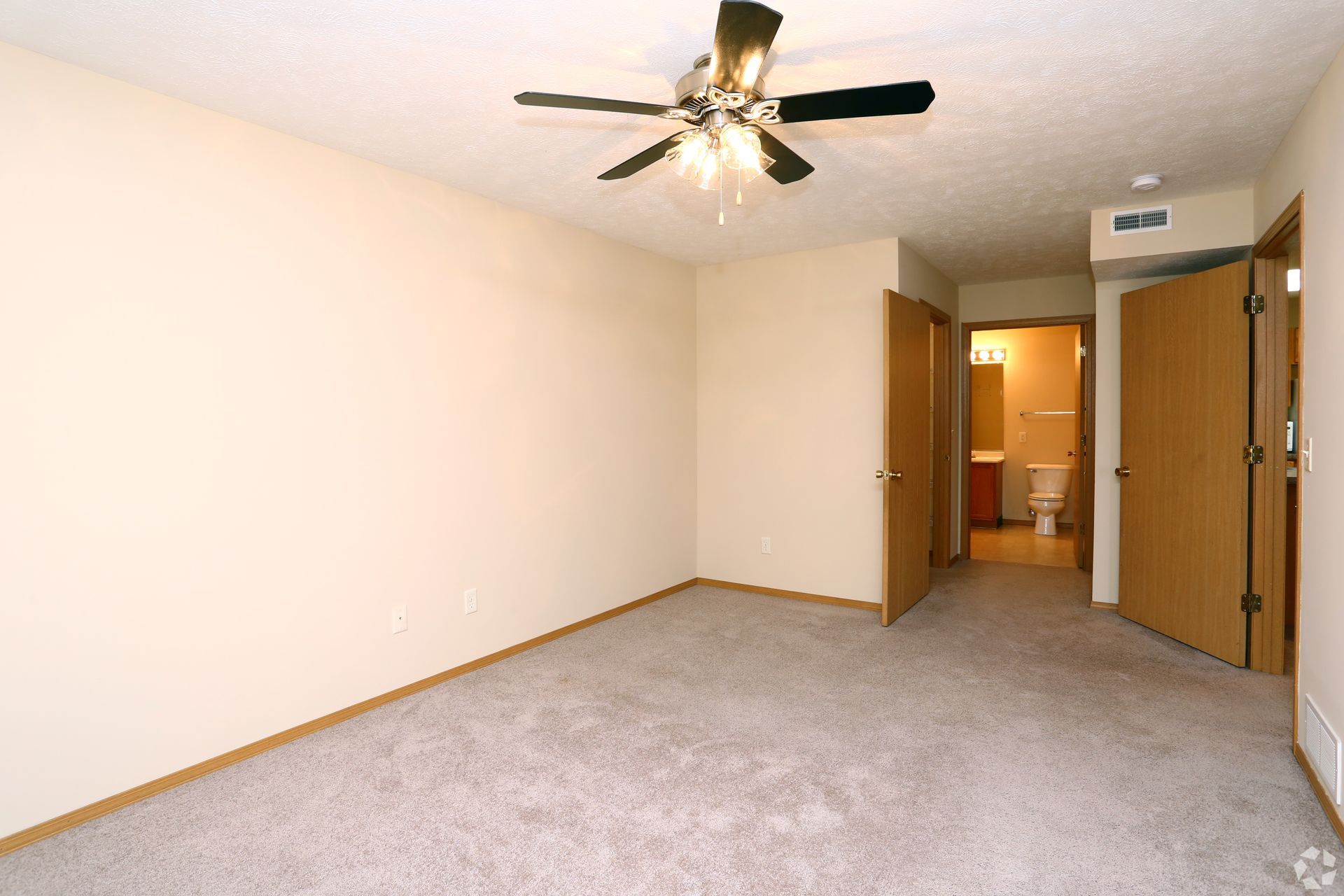Empty bedroom with tan walls, carpet, ceiling fan, and open doorways.