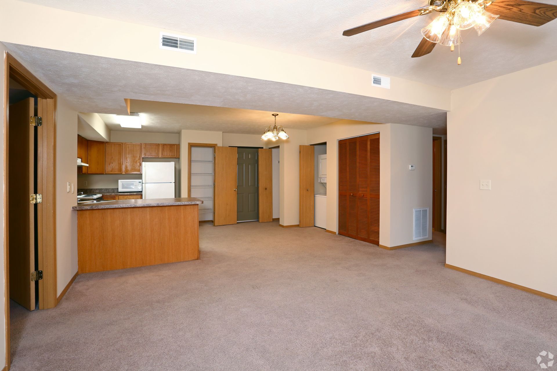 Open-plan living space with kitchen, dining area, and doors. Carpeted floor, tan walls, wooden trim and cabinets.