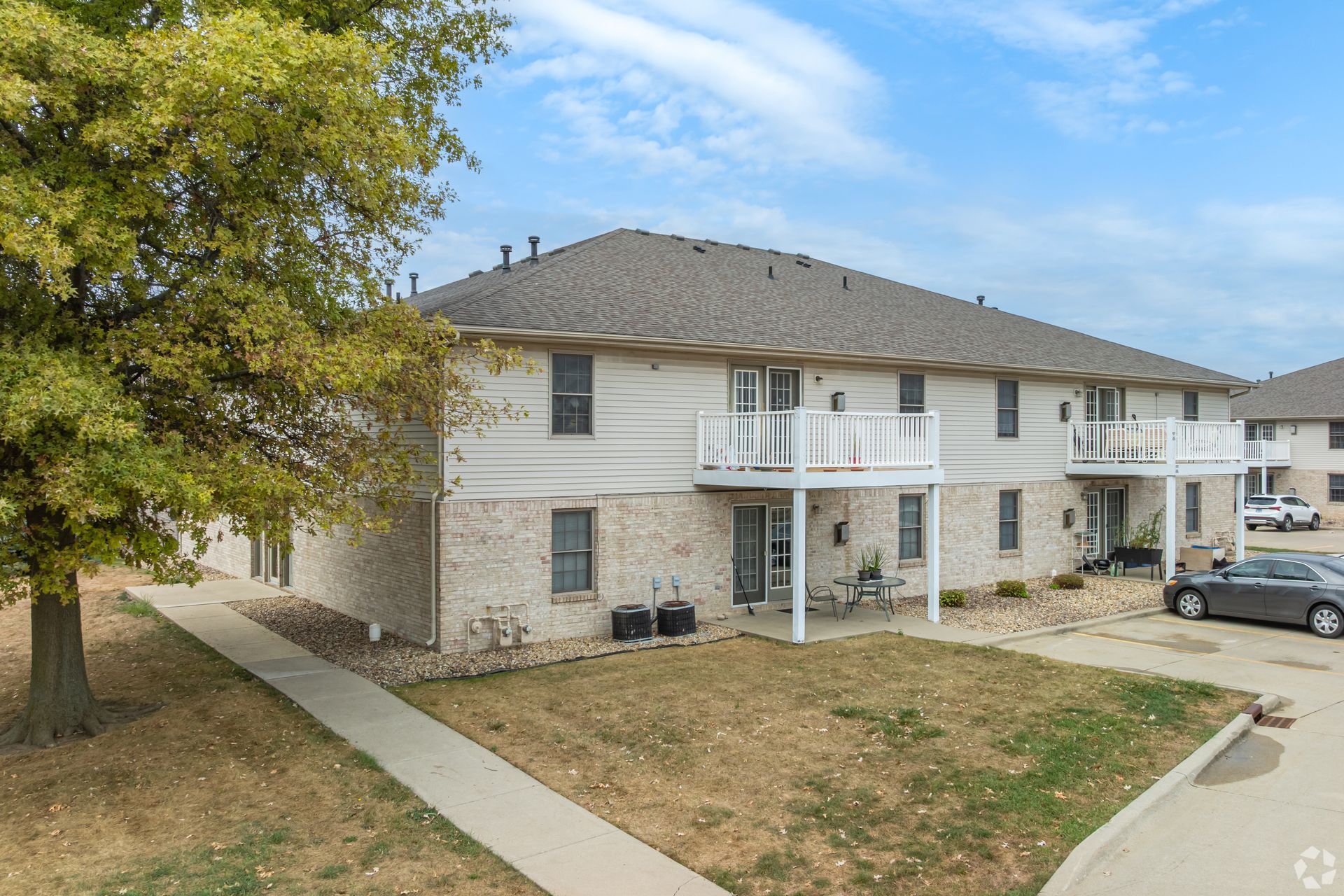 Two-story apartment building with balconies, beige siding, brick accents, and a tree in front.
