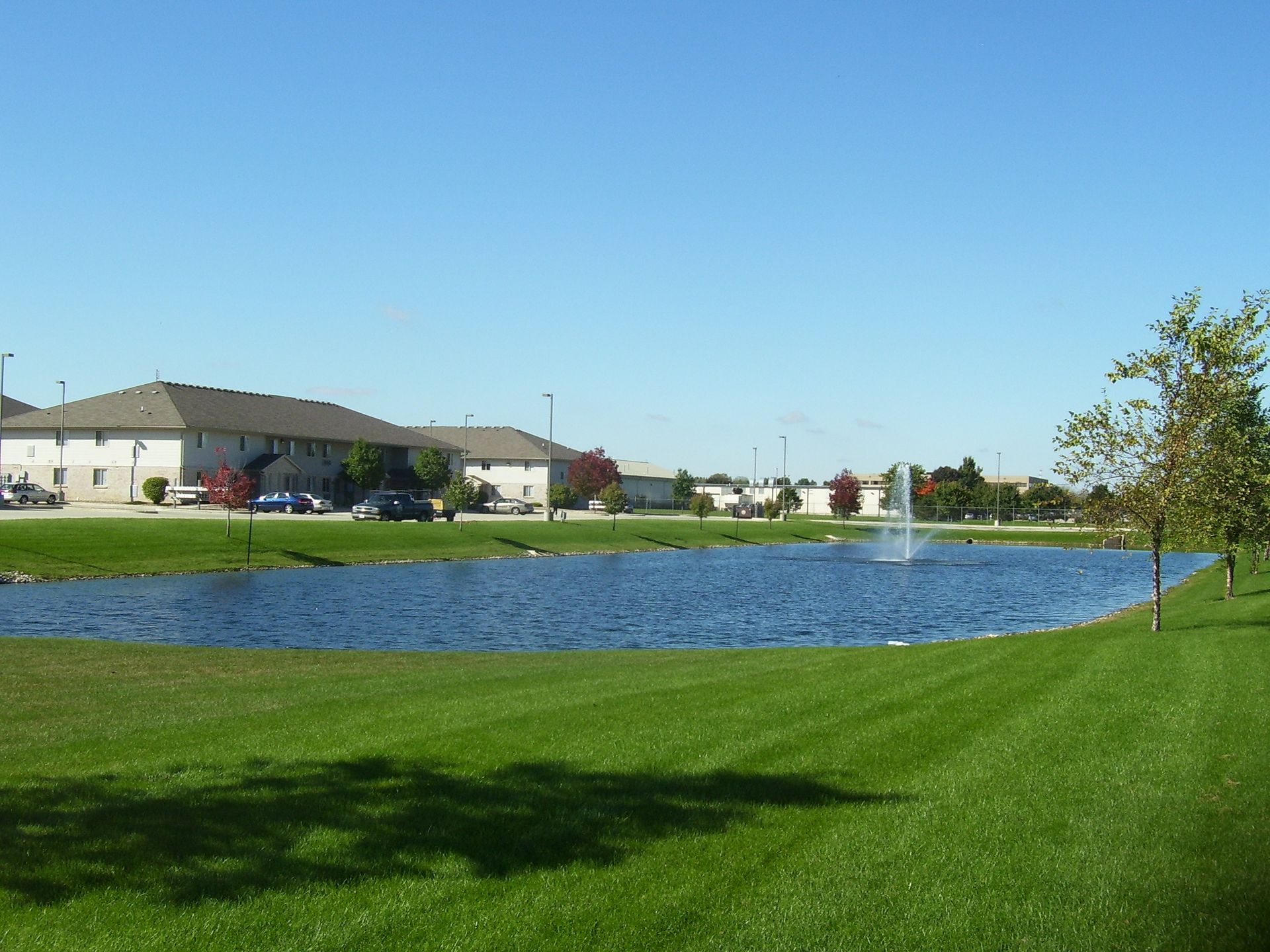 A pond with a fountain, green grass, and low buildings under a blue sky.
