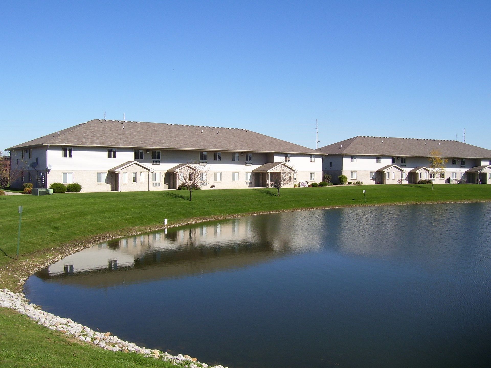 Two-story apartment buildings with tan roofs and white siding next to a pond under a blue sky.