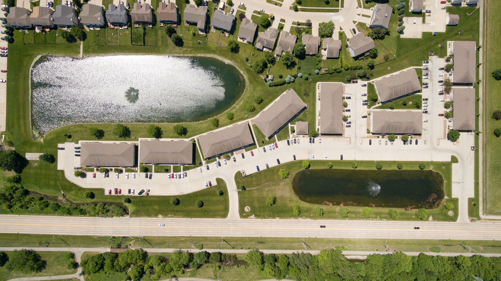 Aerial view of apartment complex with two ponds and a road. Brown rooftops, green grass.