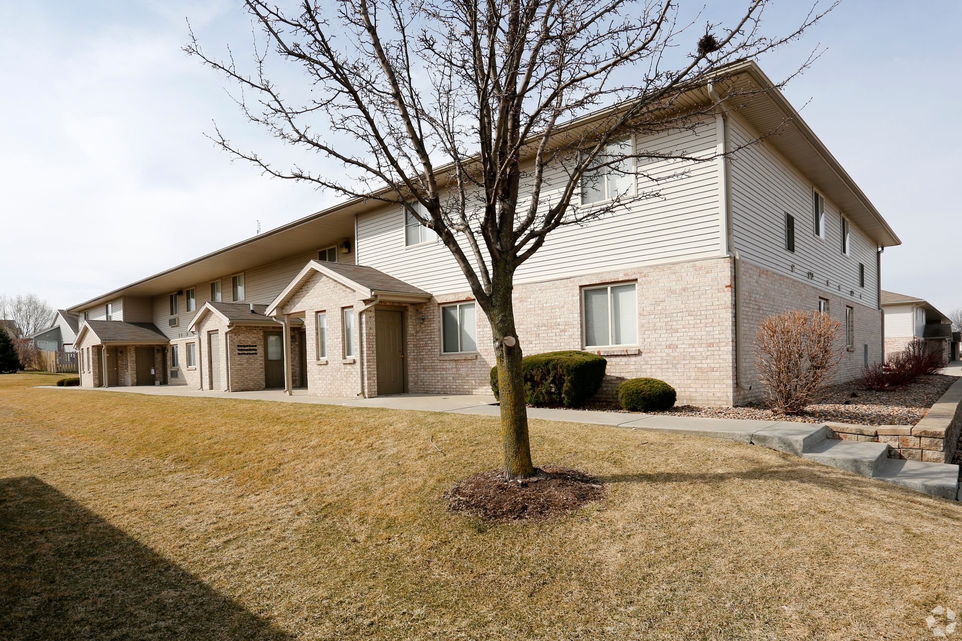 Apartment building with brick accents and brown, dry grass lawn under a mostly cloudy sky.