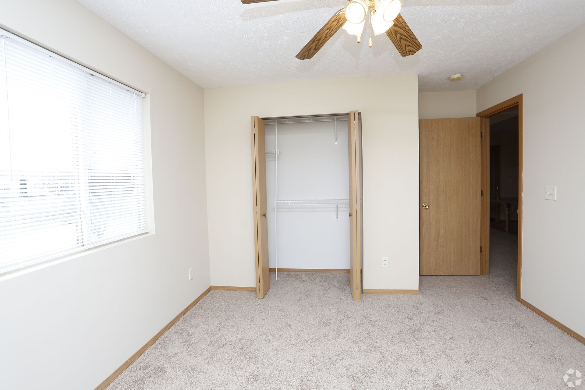 Empty bedroom with closet, window with blinds, and ceiling fan; neutral colors.