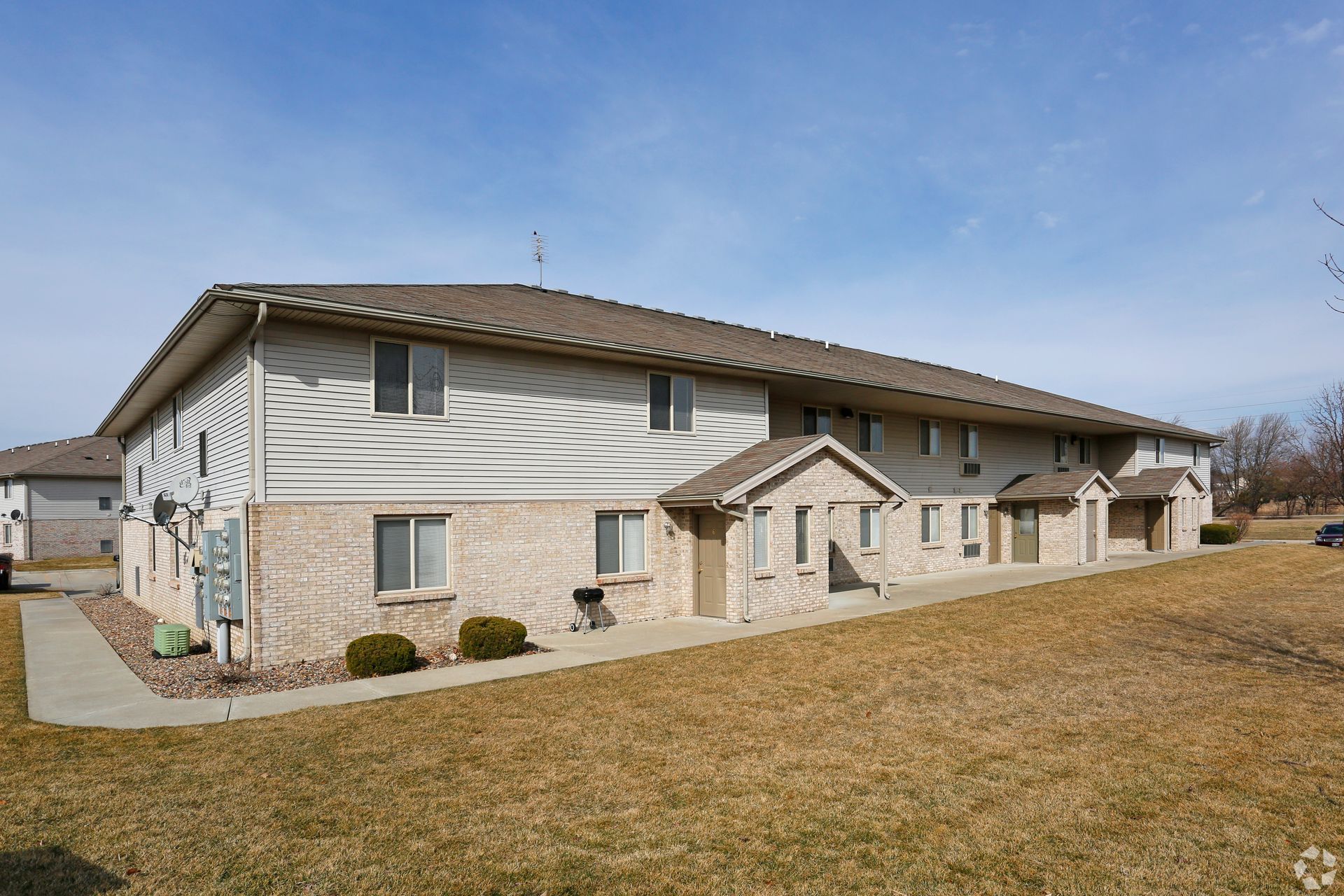 Two-story beige apartment building with brick accents and brown roof under a blue sky.