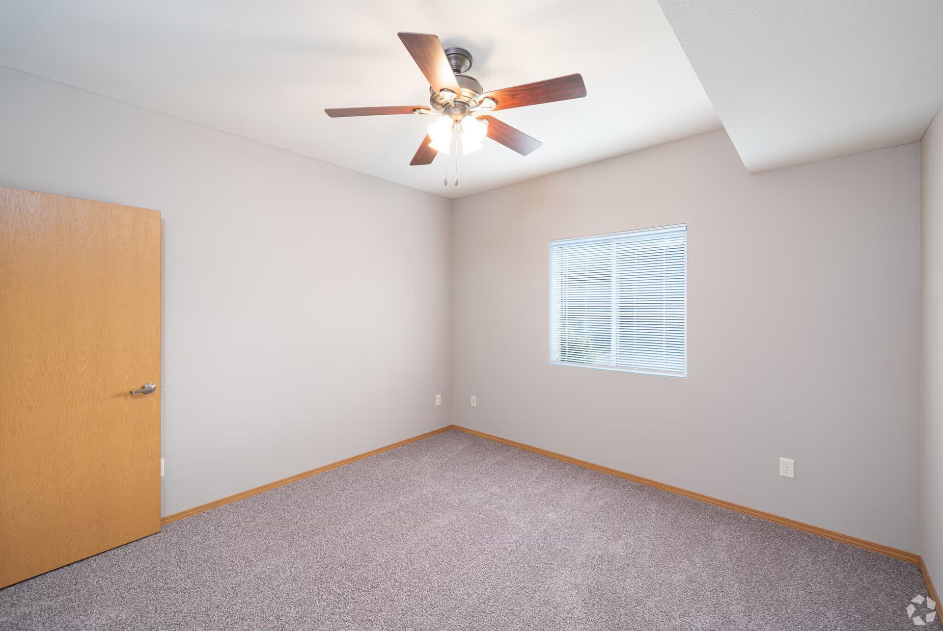 Empty bedroom with beige walls, carpet, window with blinds, and ceiling fan.