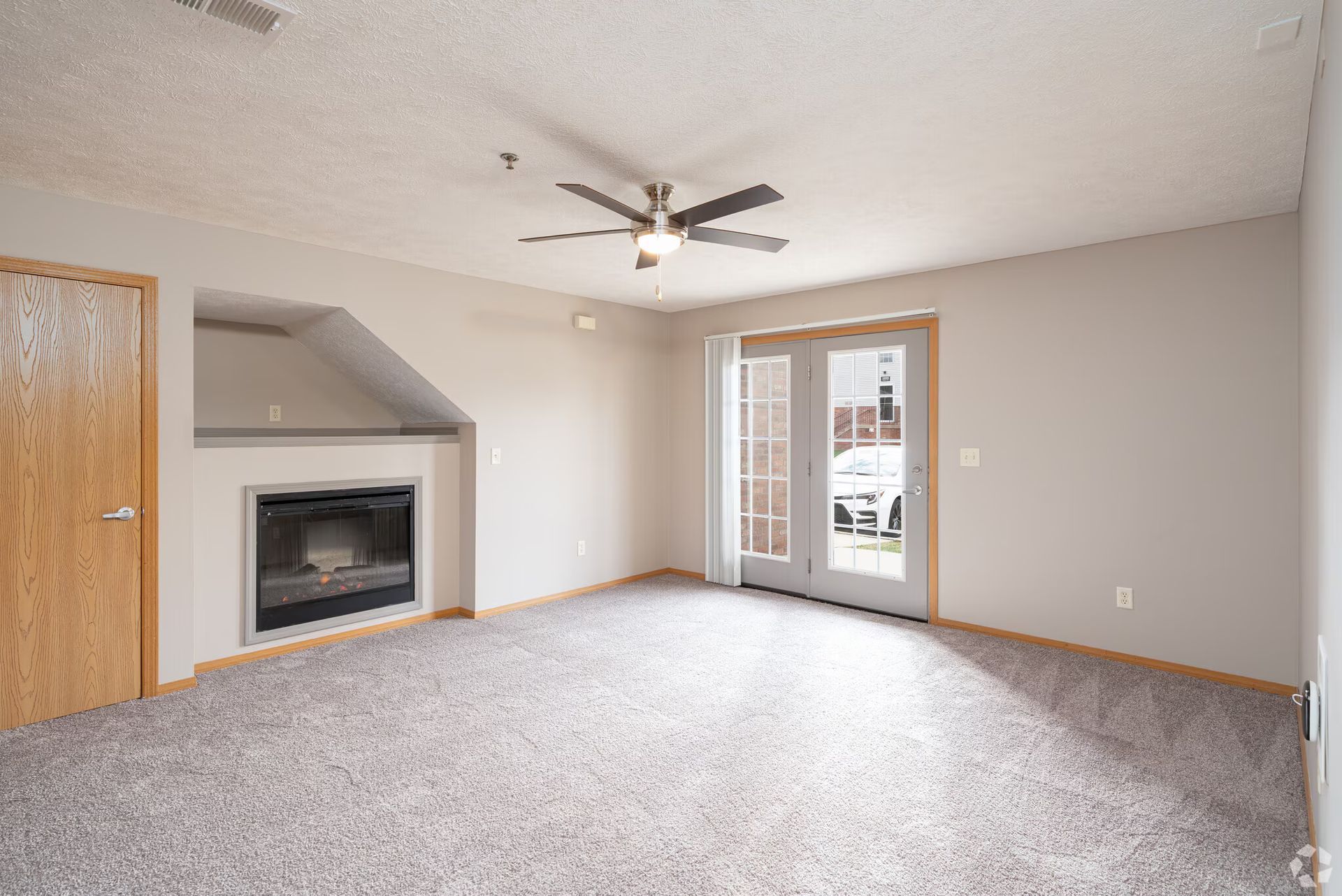 Empty living room with fireplace, French doors, ceiling fan, and gray carpet.