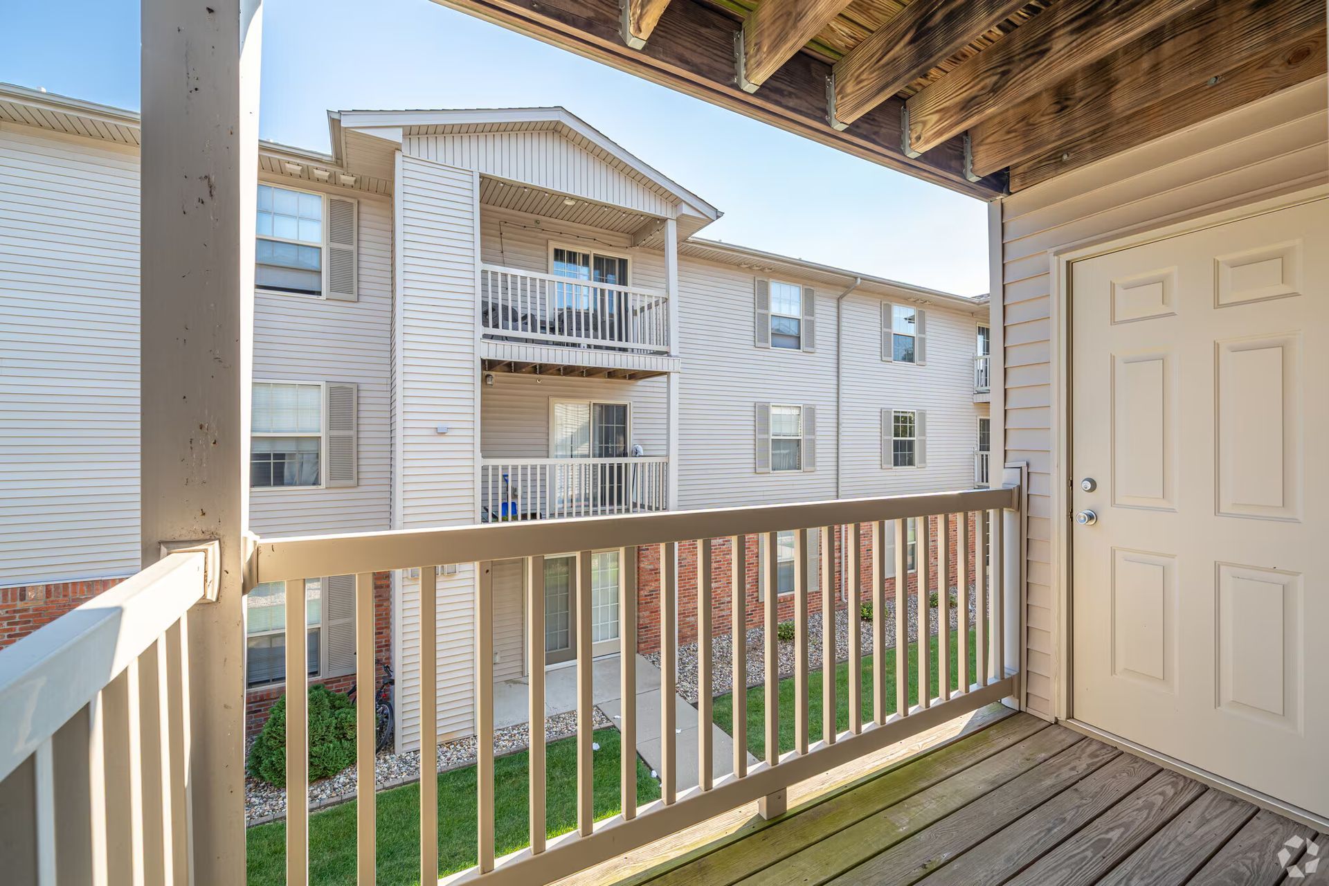 View from a balcony of an apartment building. Beige siding, white railing, and a white door are visible.