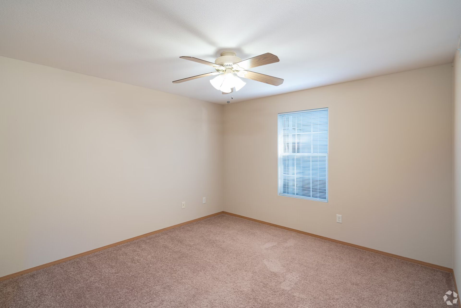 Empty bedroom with tan walls, brown carpet, ceiling fan, and window with blinds.