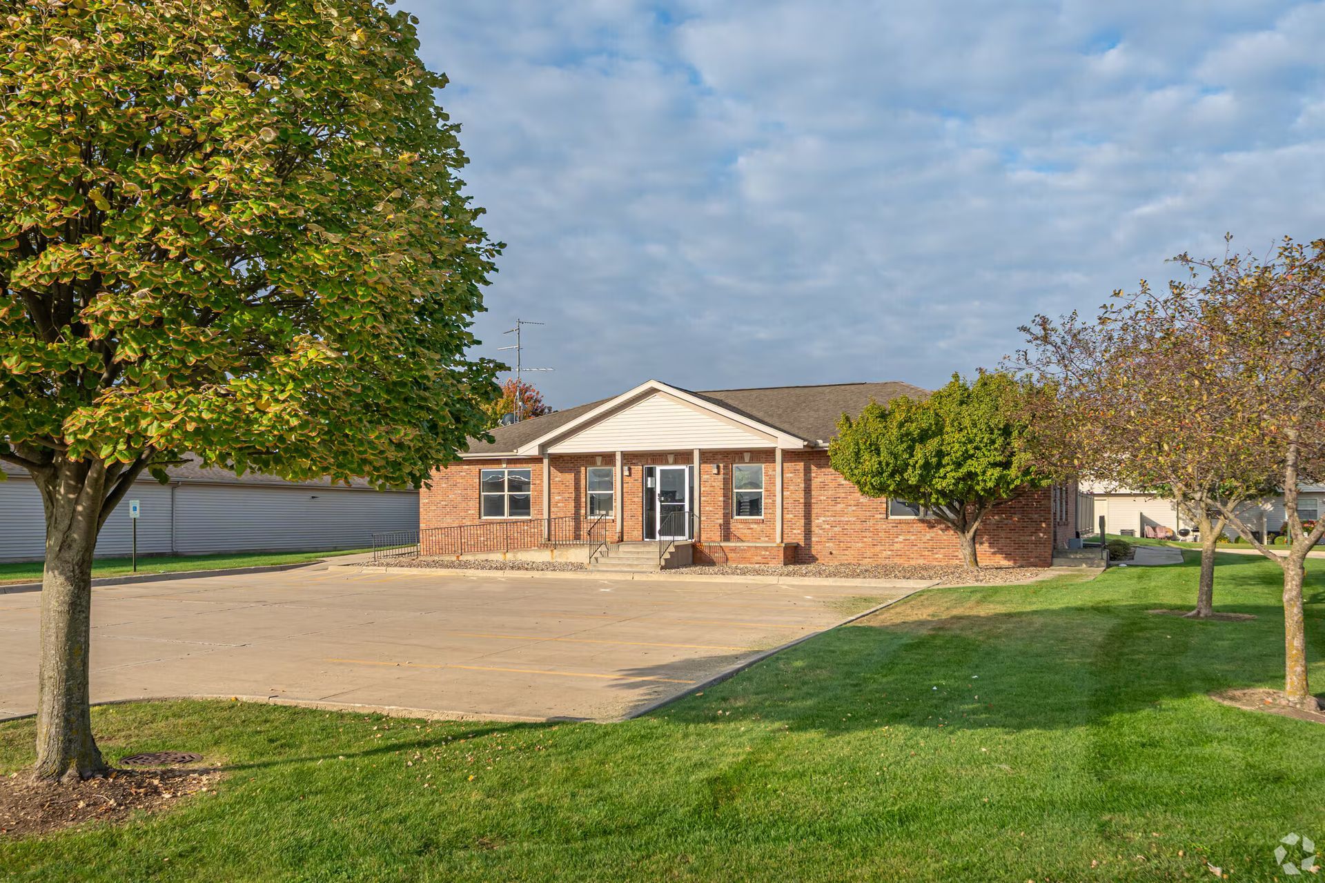 Brick building with a light-colored roof, surrounded by green grass and trees, and a paved area in front.