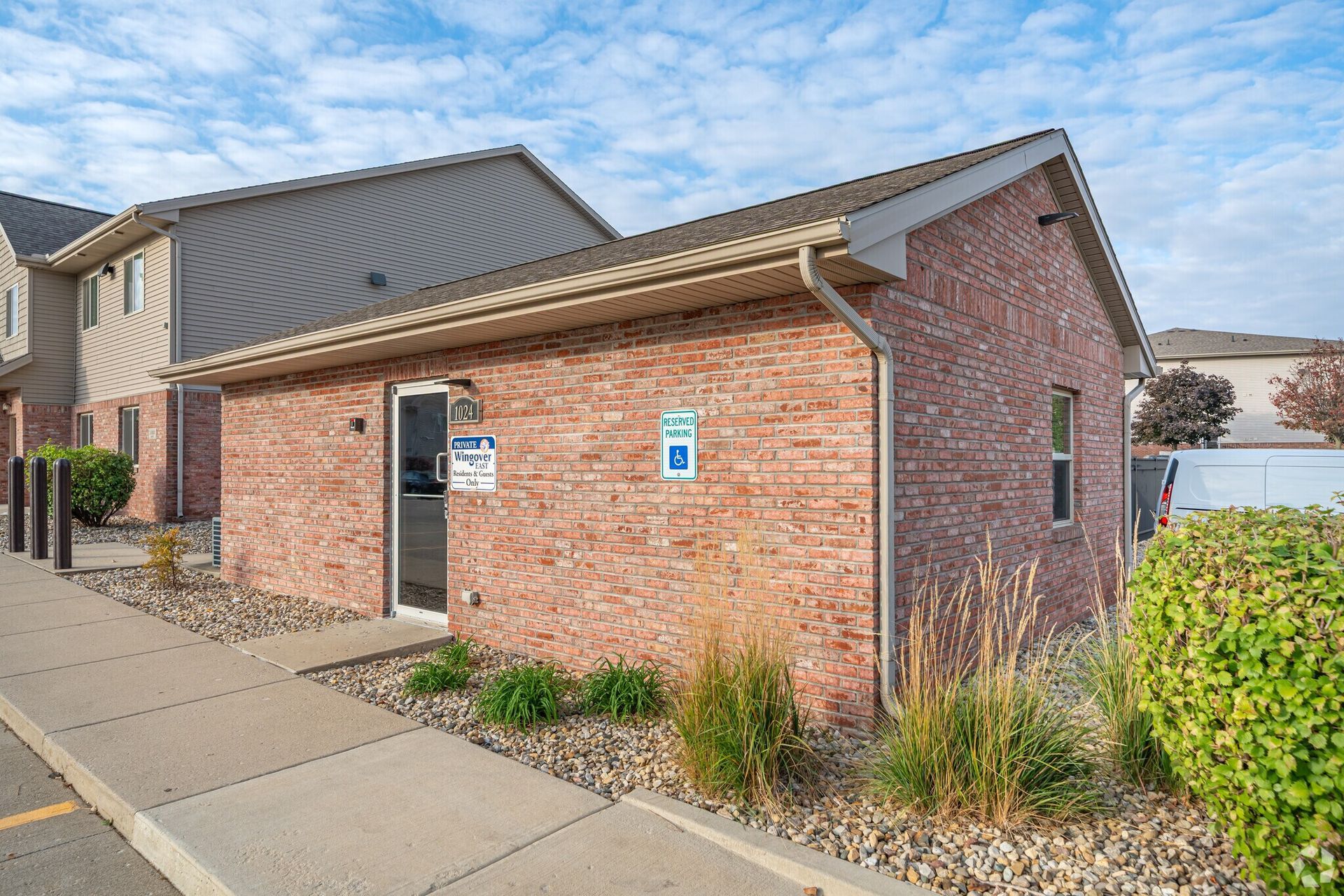 Brick building with a door, window, and small plants. Brown roof and sidewalk. Cloudy sky.