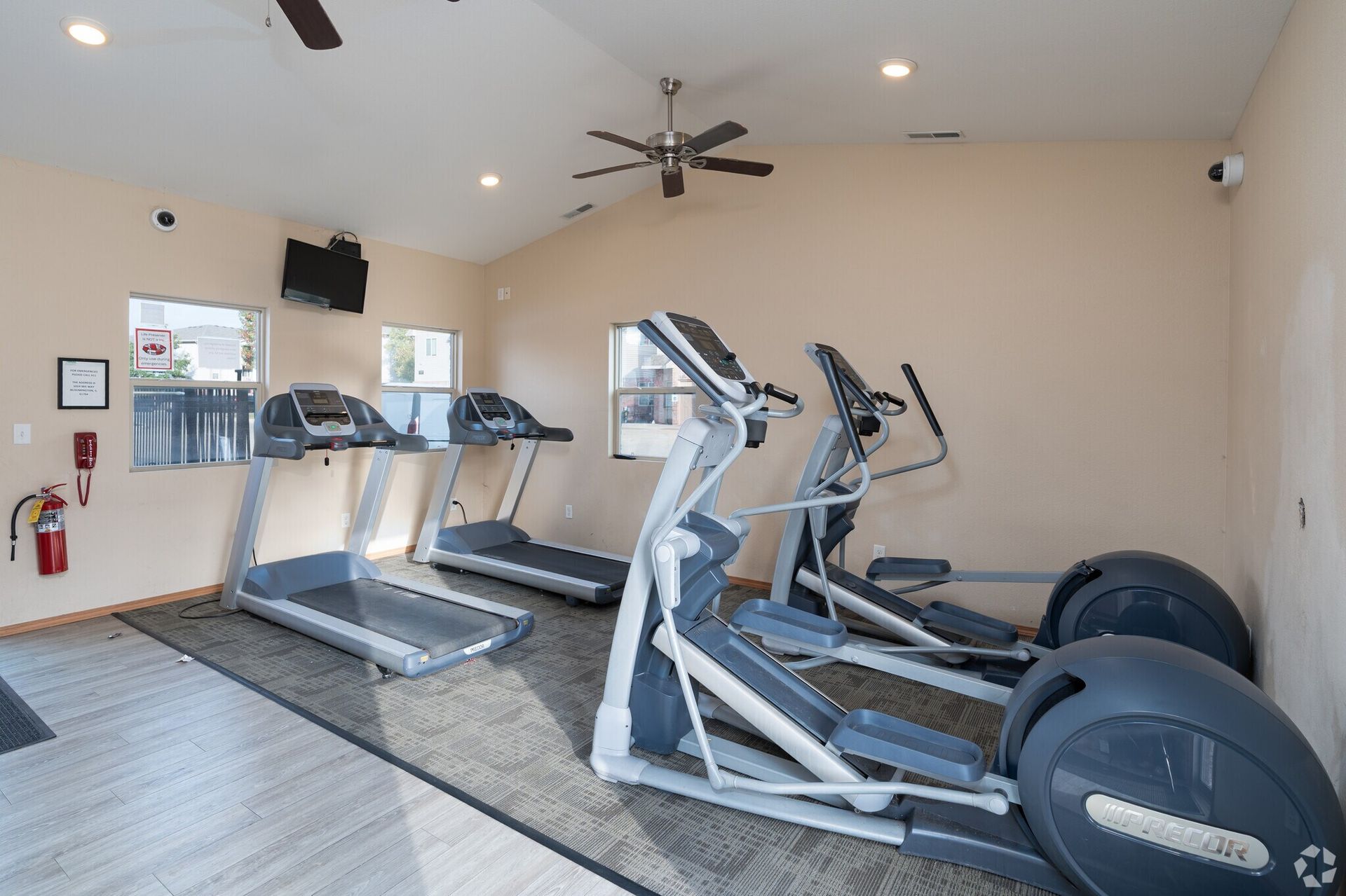 Gym interior with treadmills, elliptical, TV, windows, and ceiling fan. Beige walls, gray carpet.