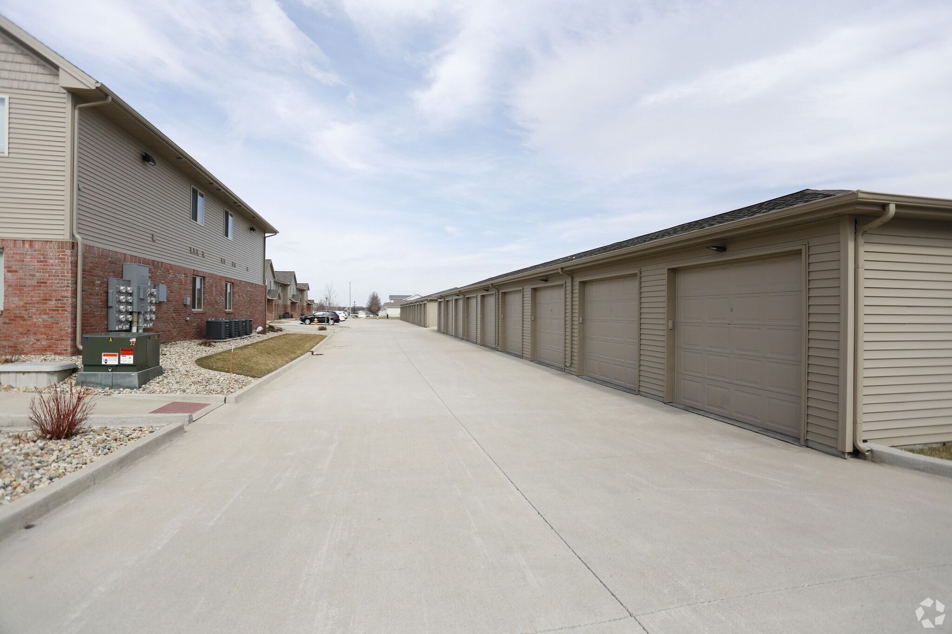 Row of attached garages with a residential building on the left and a concrete driveway under a cloudy sky.