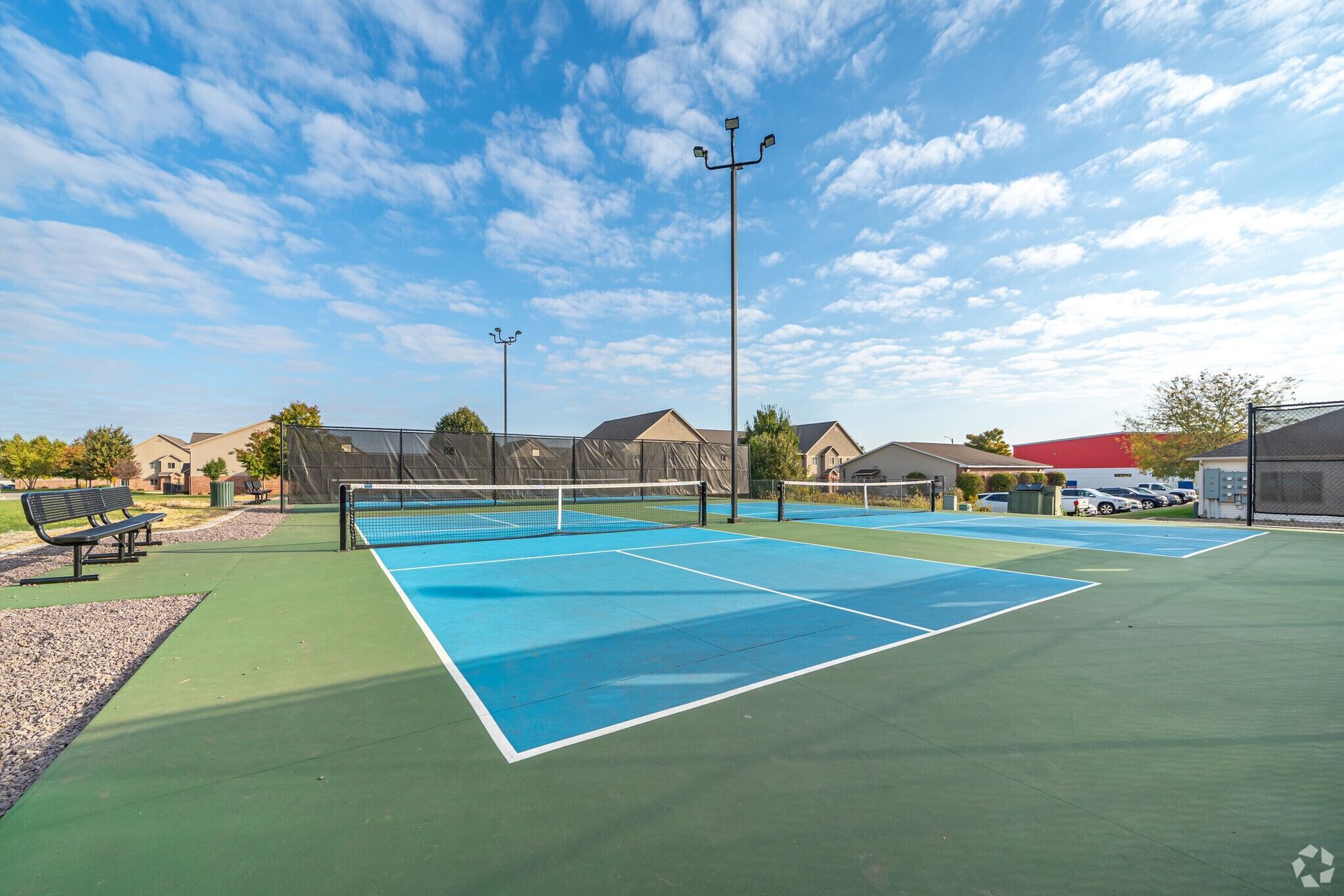 Pickleball court with blue and green surfaces, net, and light poles. Houses in the background under a blue sky.