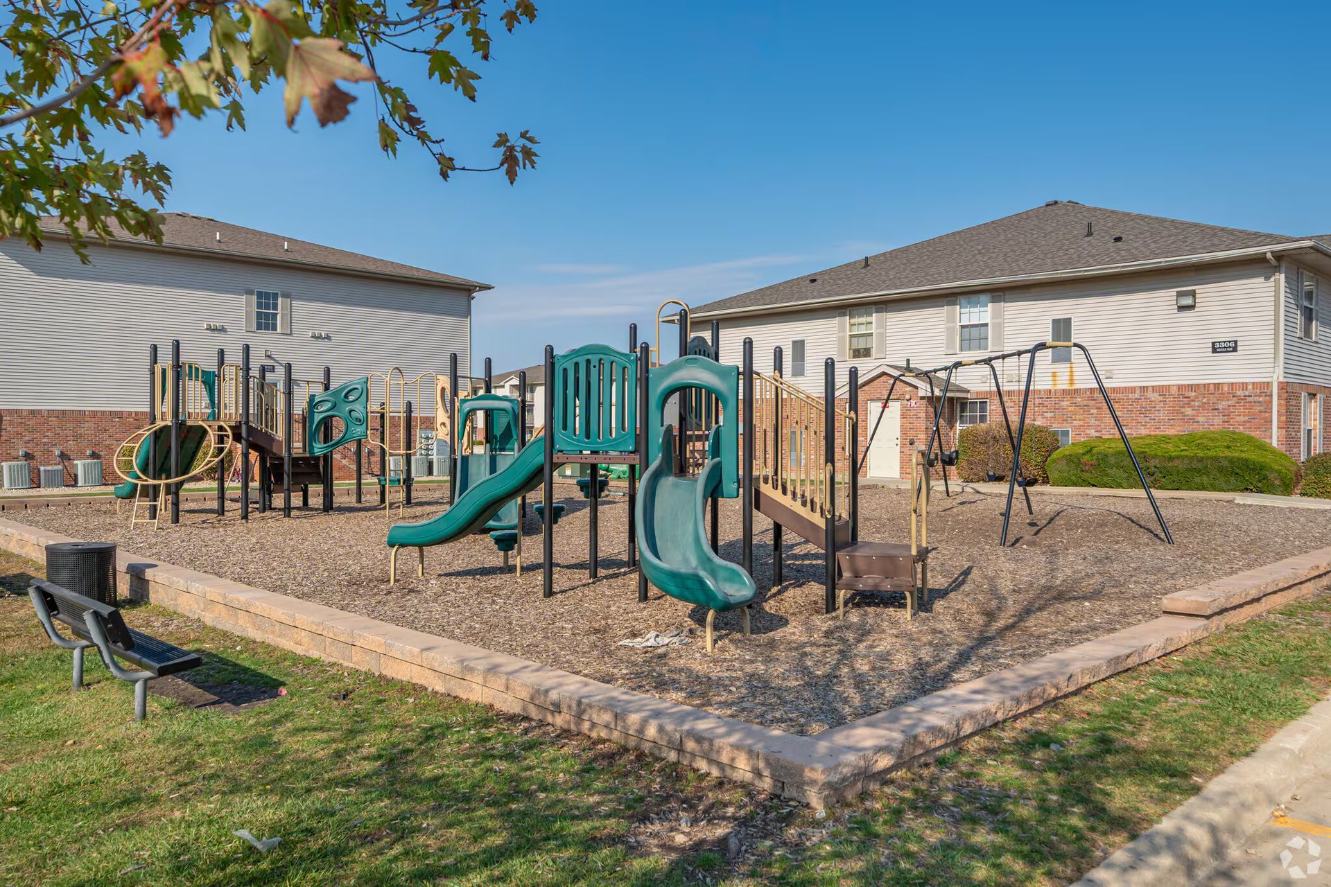 Playground with green slides, swings, and climbing structures near two tan buildings under a clear blue sky.