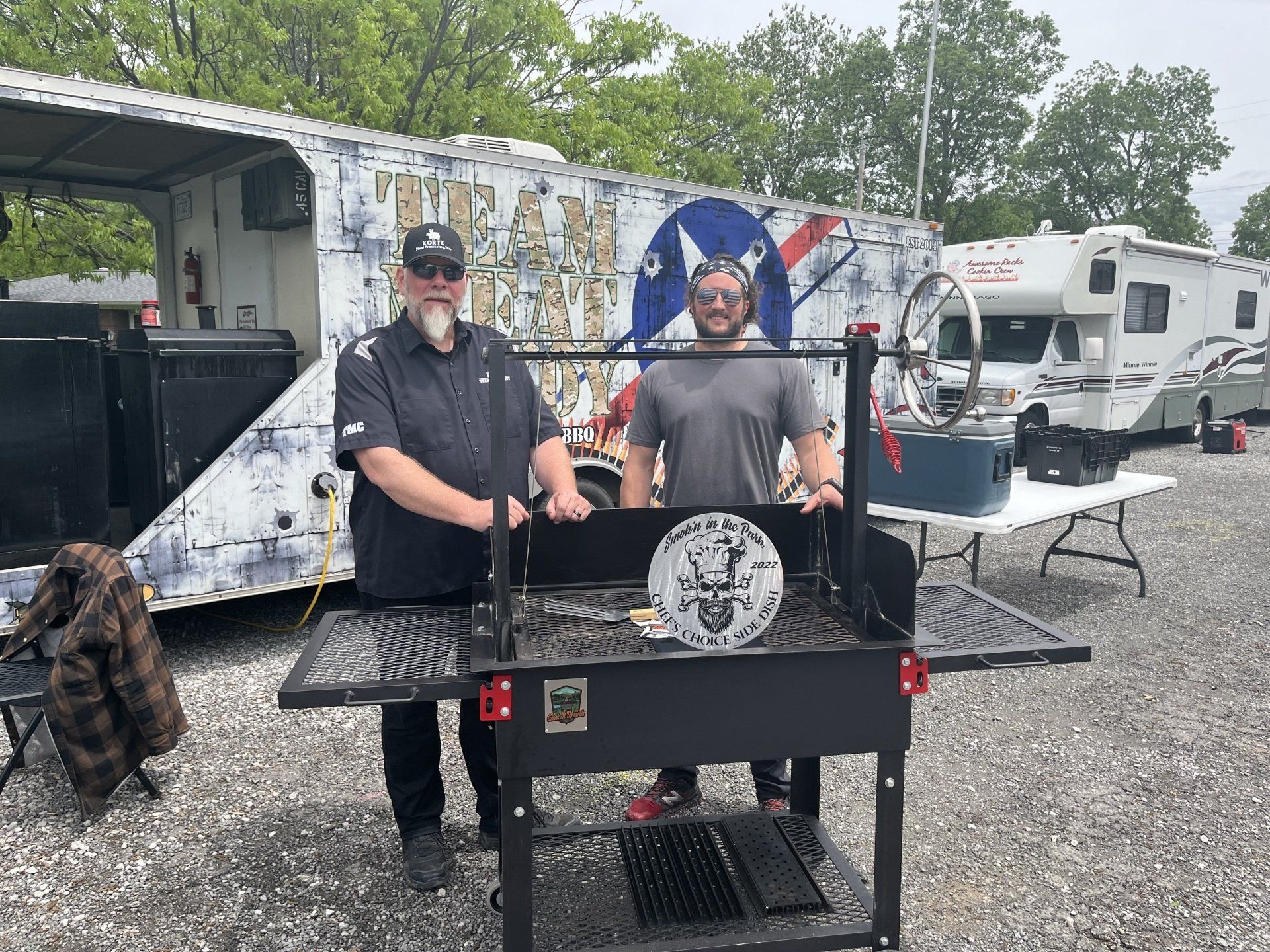 Two men are standing next to a grill in a parking lot.