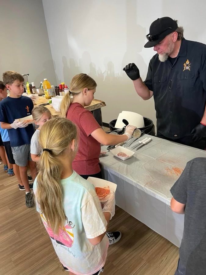 A group of children are standing around a table while a man prepares food.