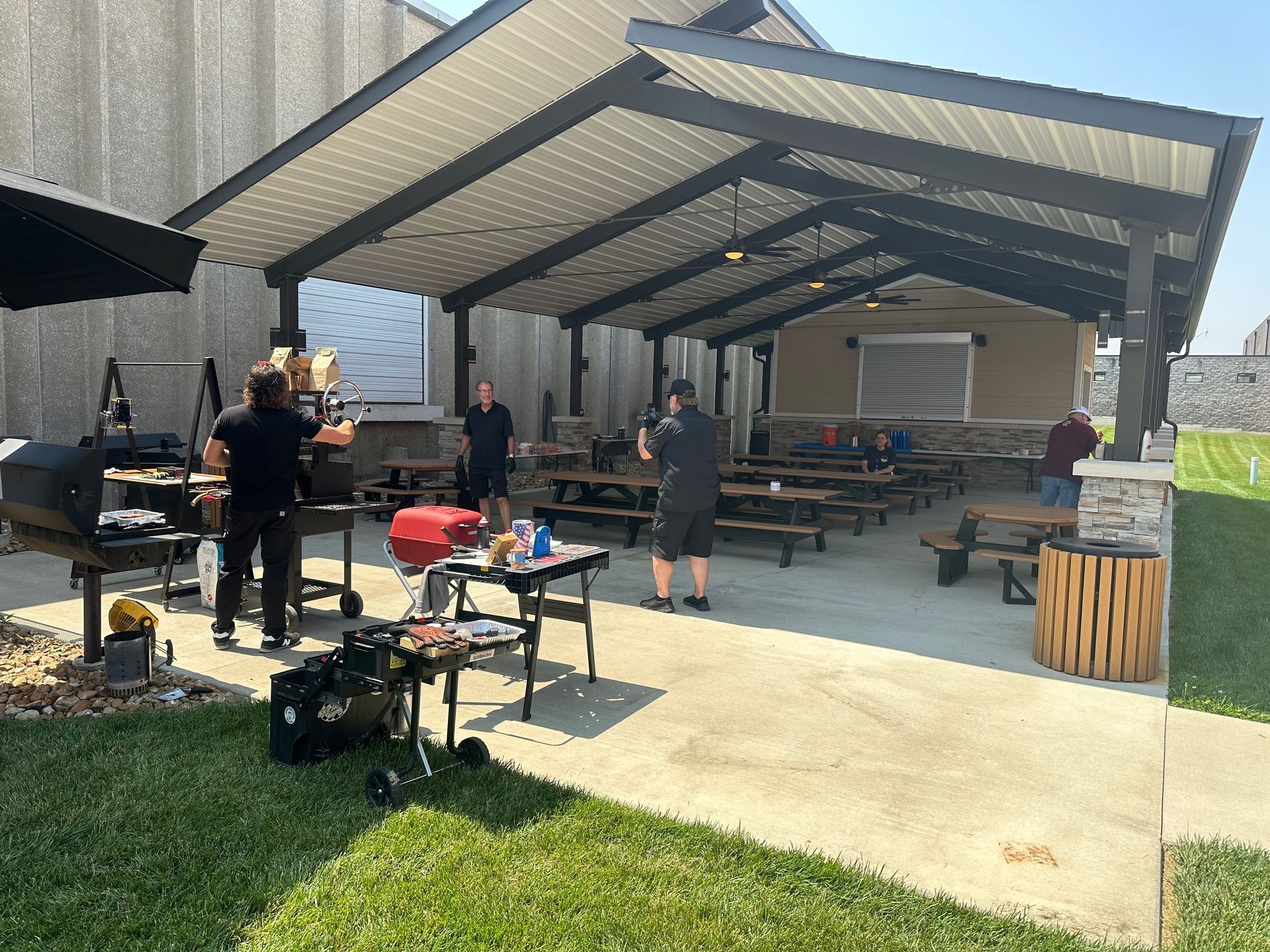 A group of people are standing under a covered area with tables and grills.