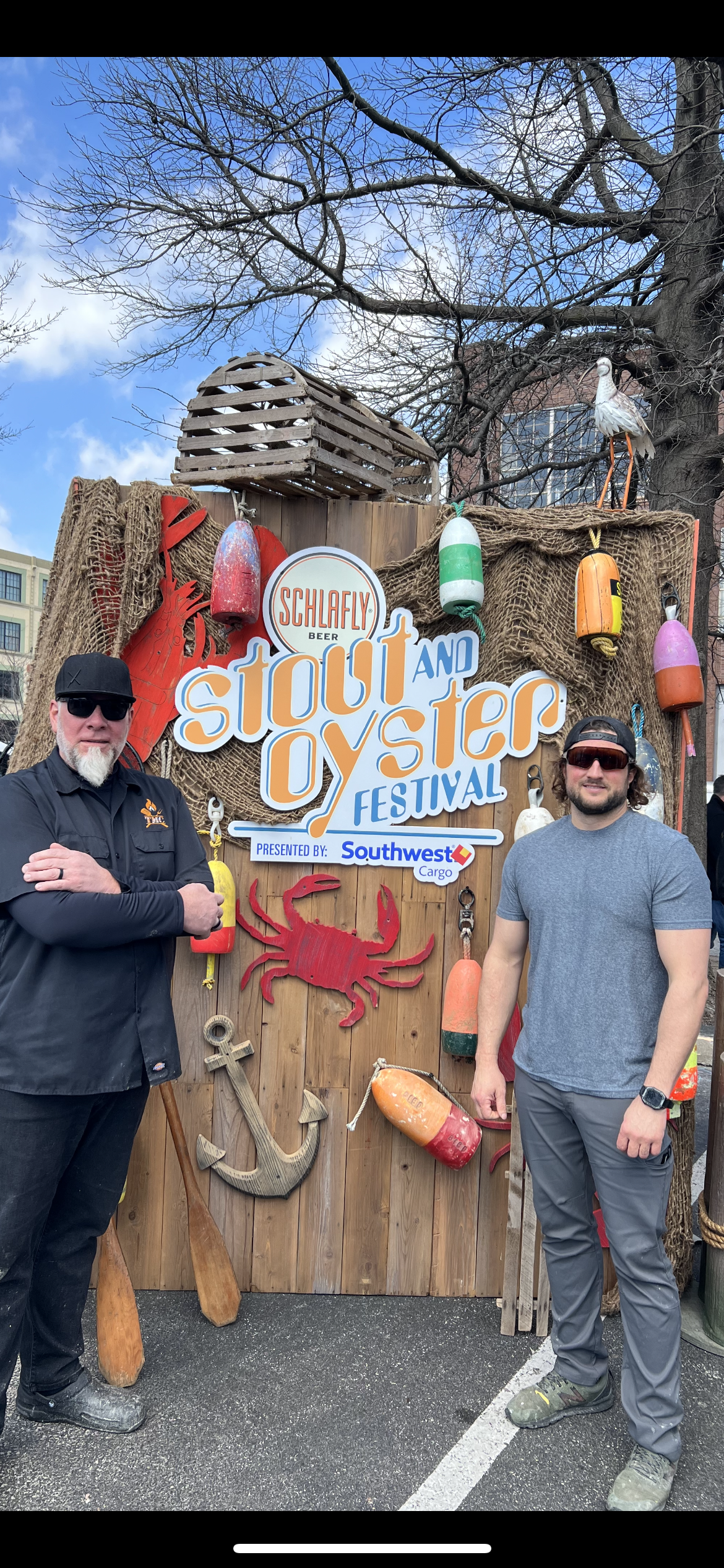 Two men are standing in front of a wooden sign.