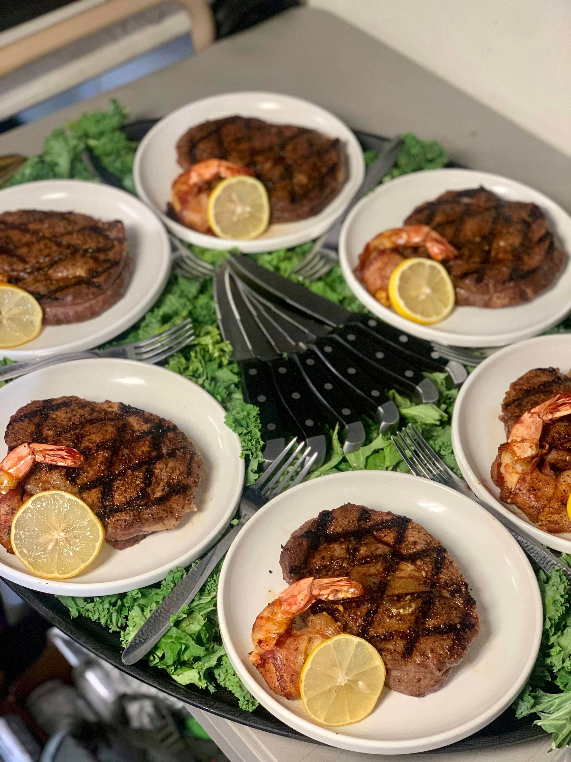 A tray of steaks and shrimp with lemon slices on a table.