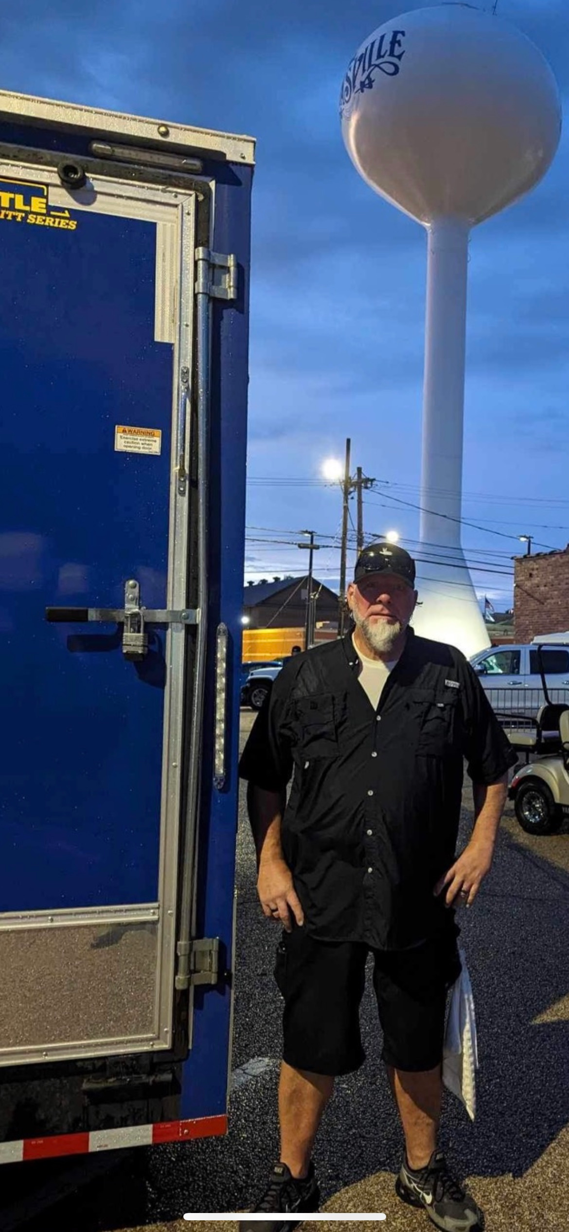 A man is standing in front of a blue truck next to a water tower.
