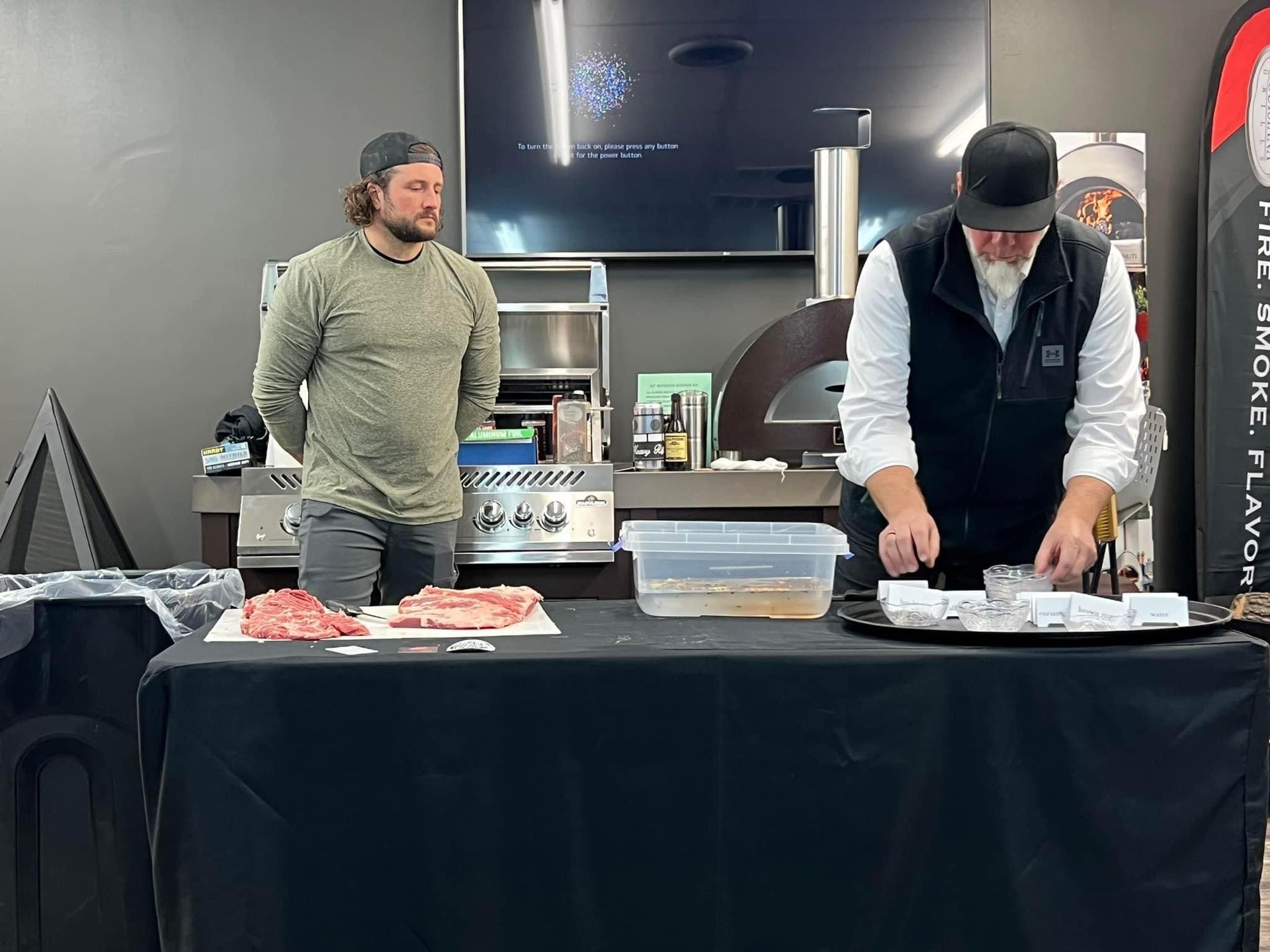 Two men are standing in front of a table with meat on it.