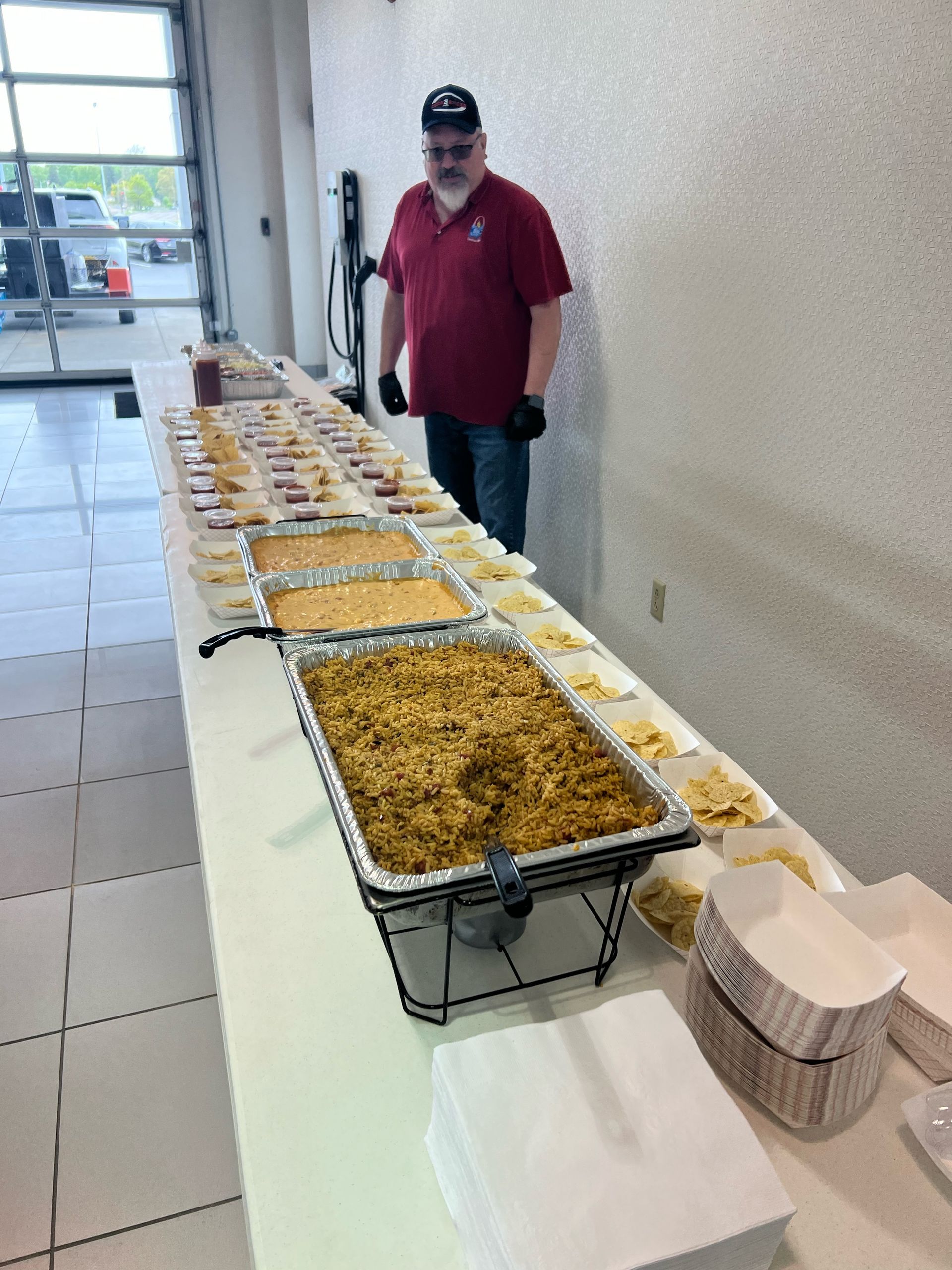 A man in a red shirt is standing in front of a long table filled with trays of food.