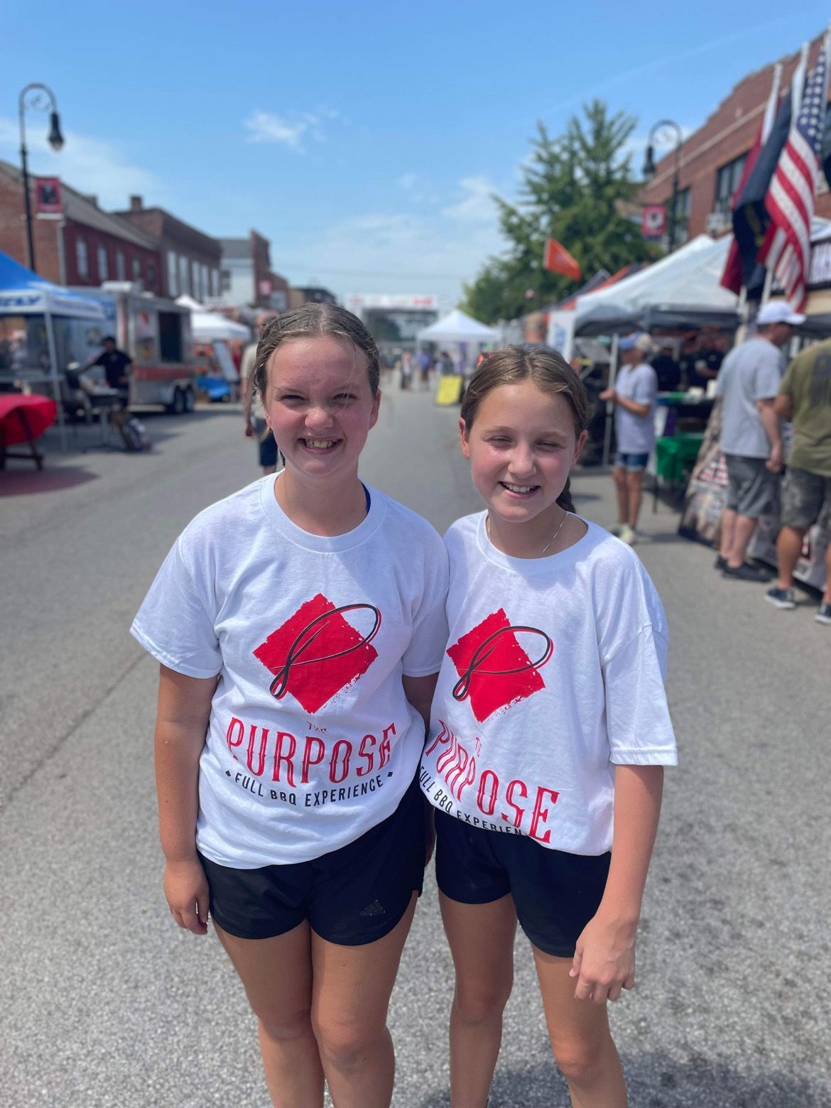 Two young girls wearing white shirts with the word purpose on them are standing next to each other on a street.