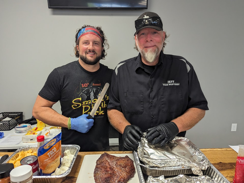 Two men are standing next to each other in front of a table with food.