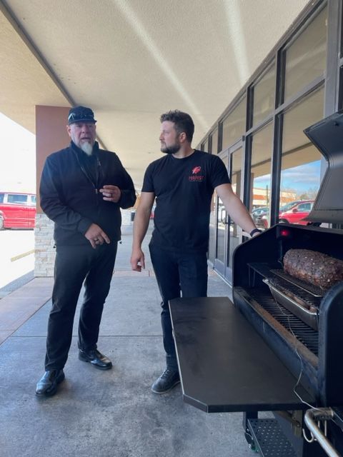 Two men standing next to a grill with a large piece of meat on it