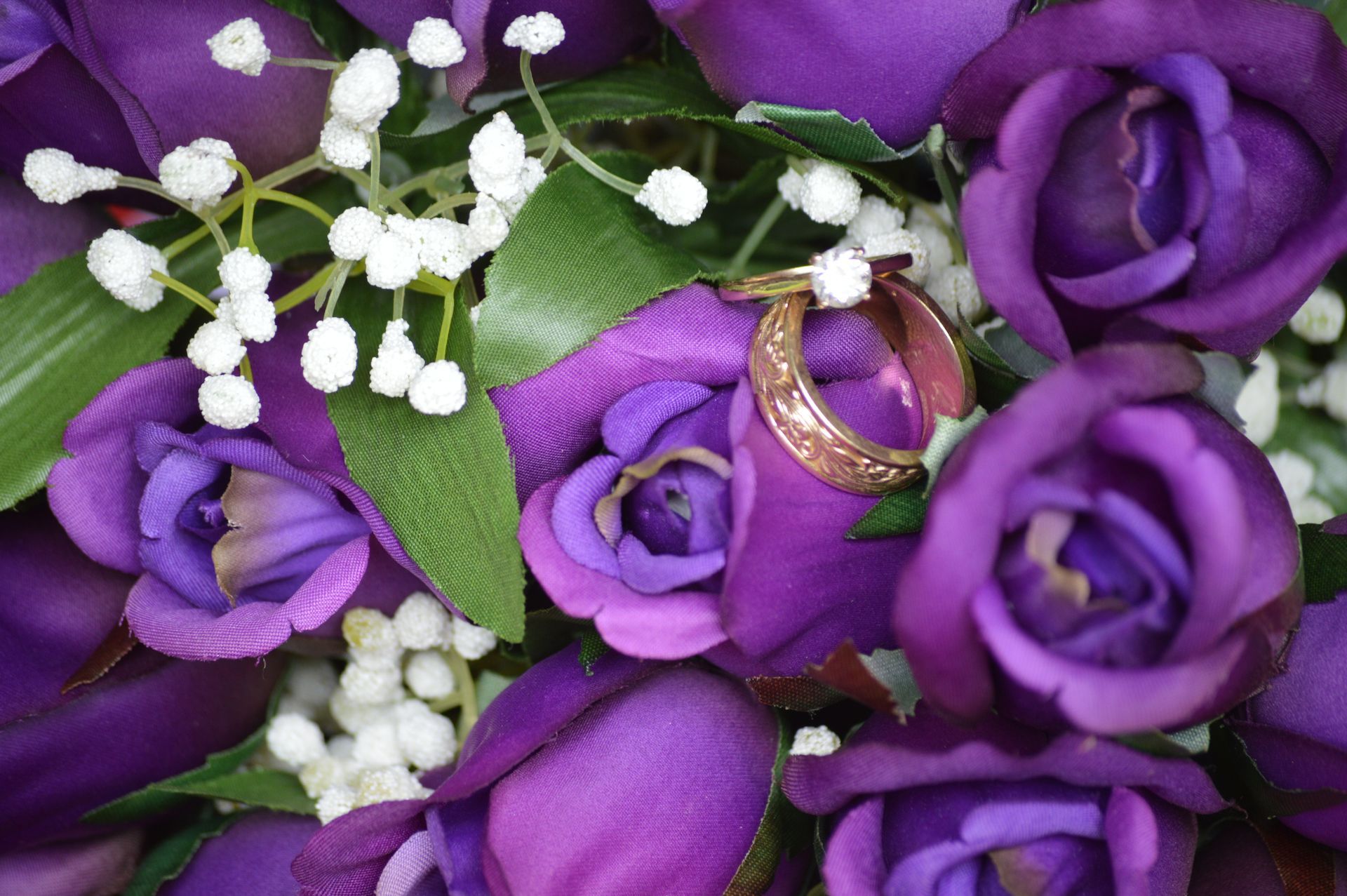 Purple roses with a gold ring nested inside; white filler flowers and green leaves.