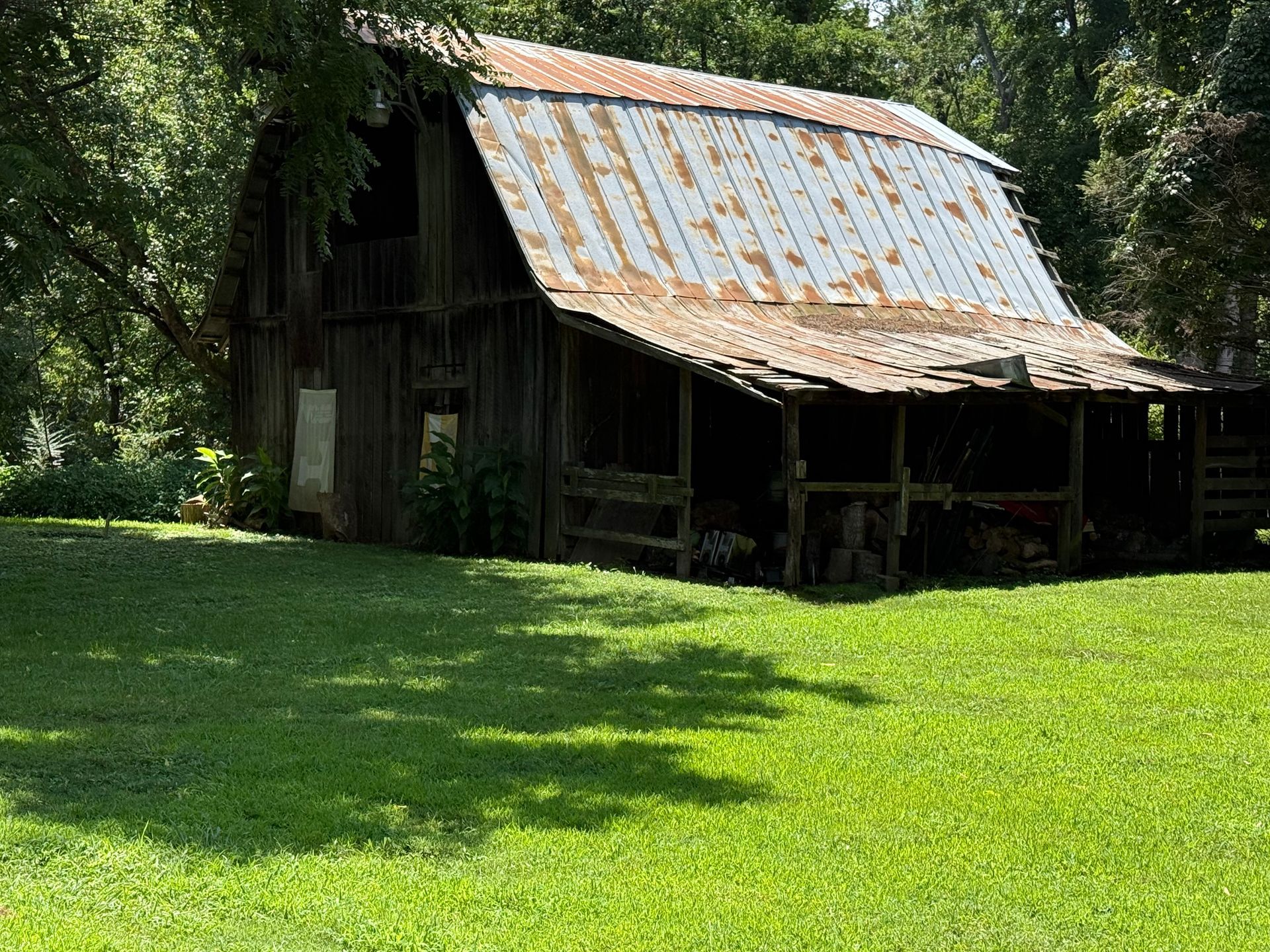 Old wooden barn with a rusty roof in a grassy field, surrounded by trees.