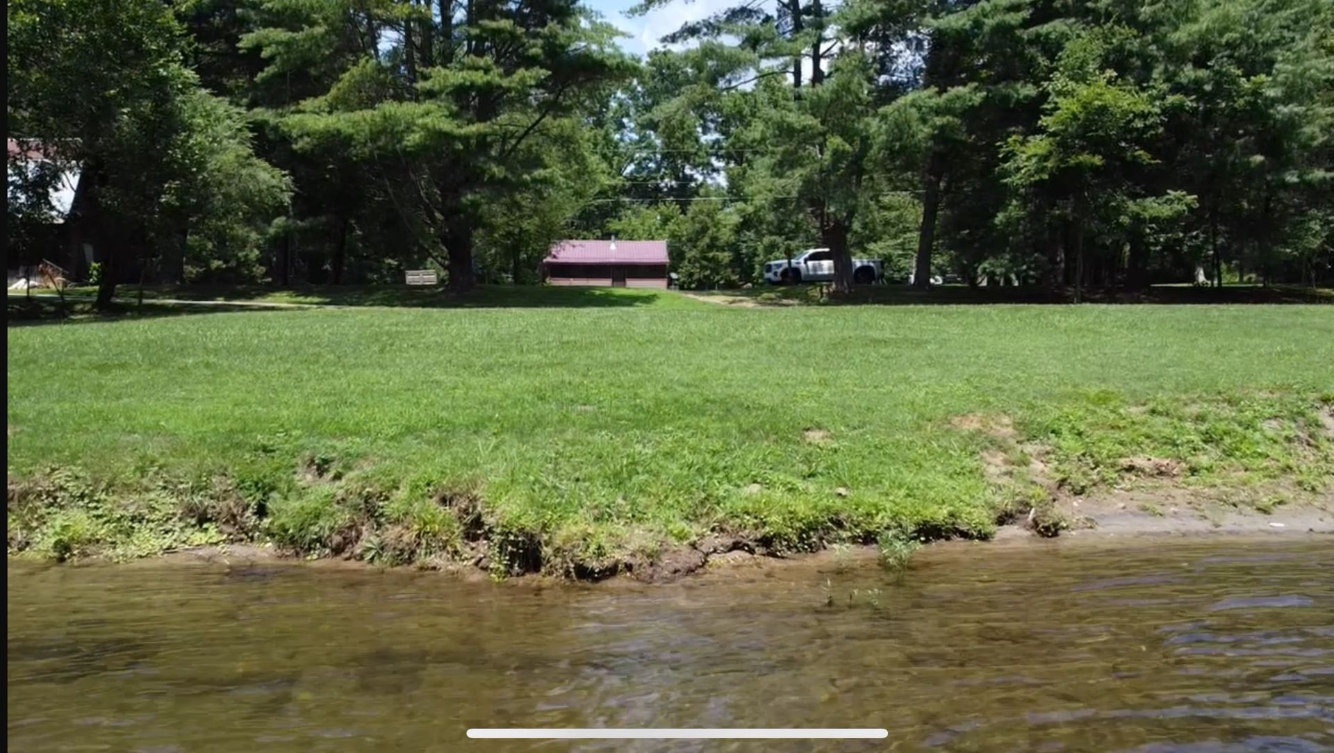 Lush green lawn bordering a body of water, with a small building and trees in the background on a sunny day.