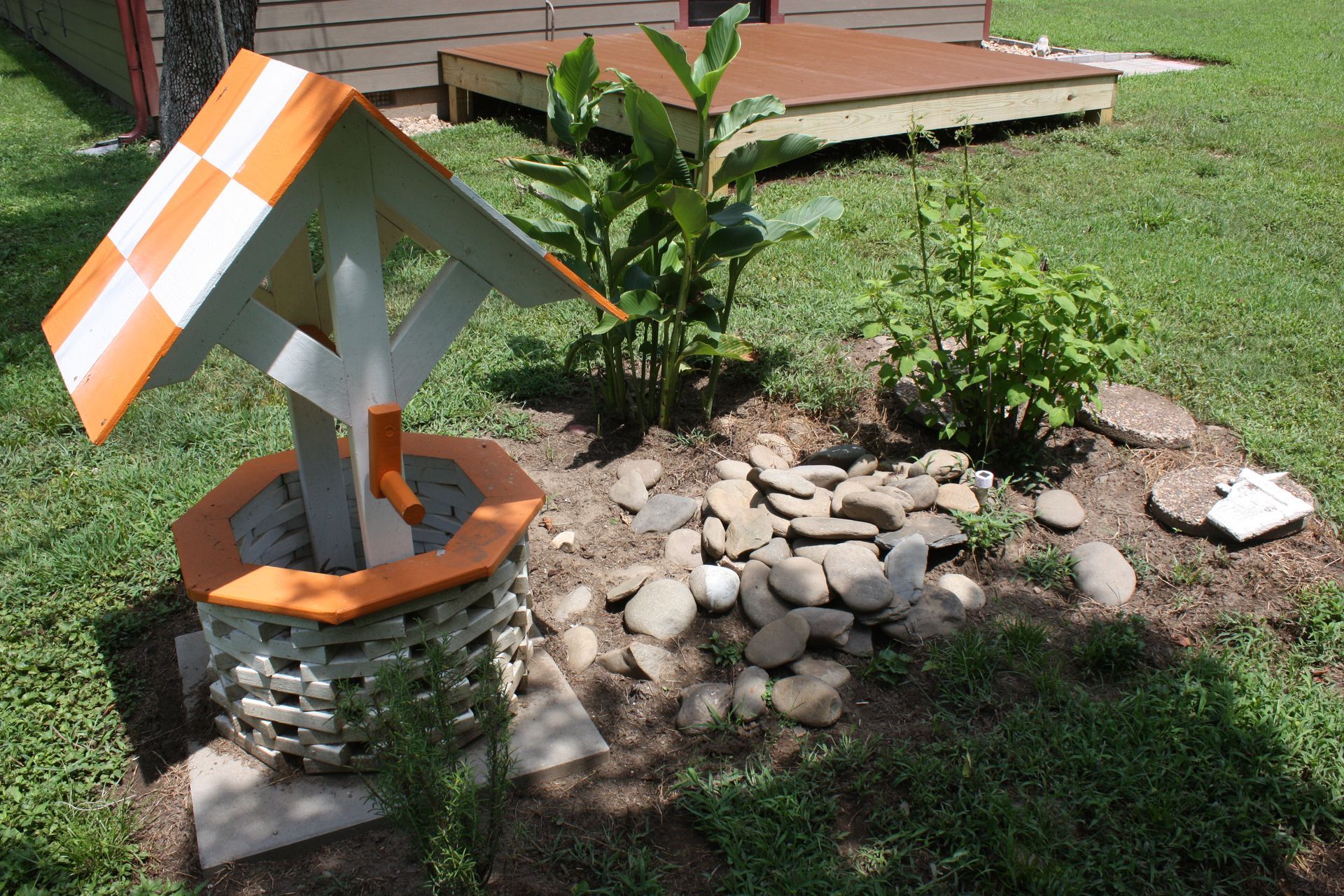 A decorative wishing well with orange and white accents sits in a garden bed with plants and rocks.