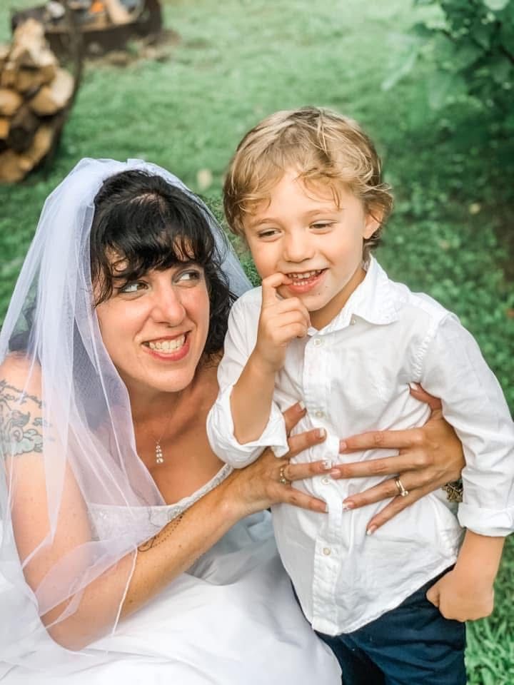 Bride in white dress and veil smiles at a young boy in a white shirt and blue pants. They are outdoors.