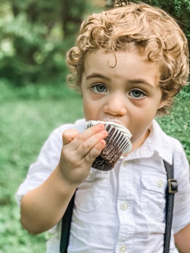 Boy with curly hair eats a cupcake outside. He wears a white shirt with suspenders.