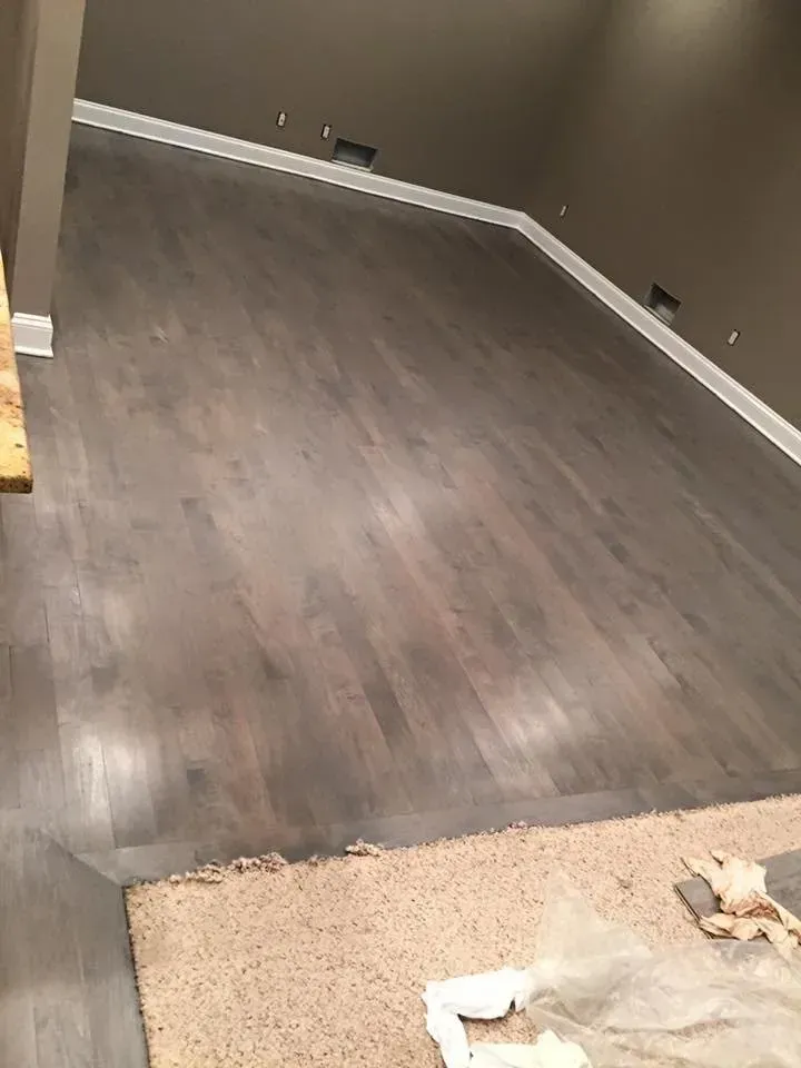 Gray wood-look flooring with white trim installed in a room with brown walls and a tan carpeted section.