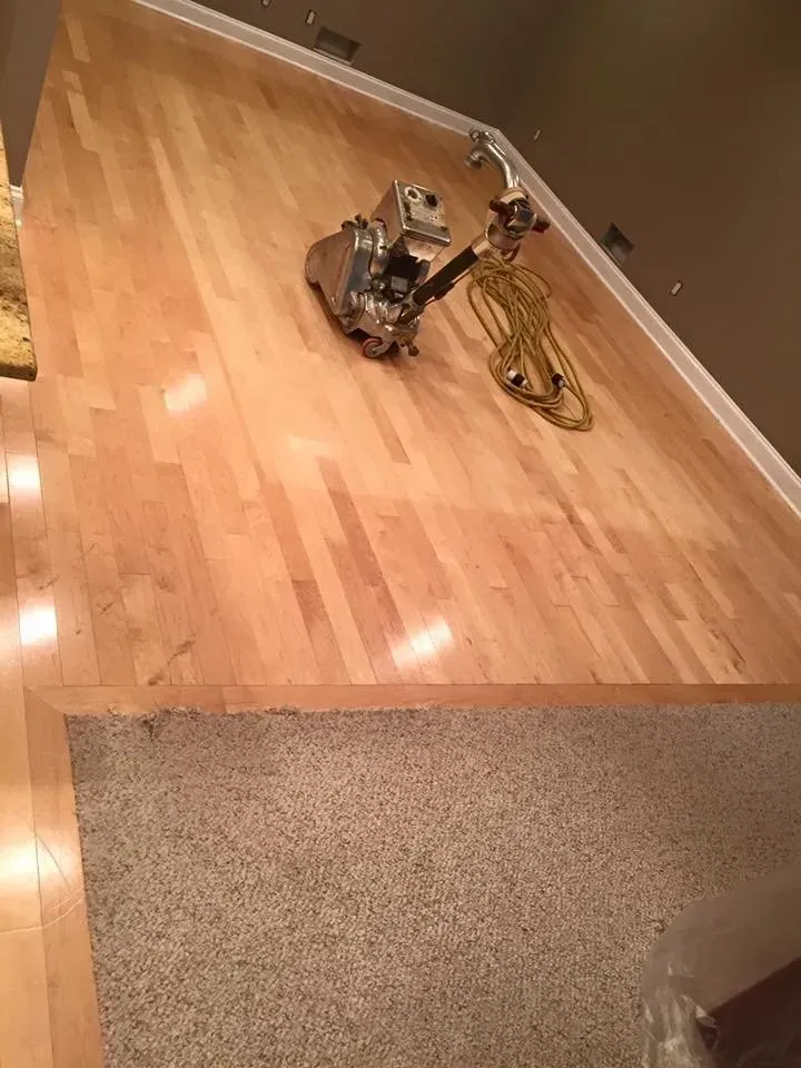 Floor sander on light wood floor next to beige carpet; neutral lighting.