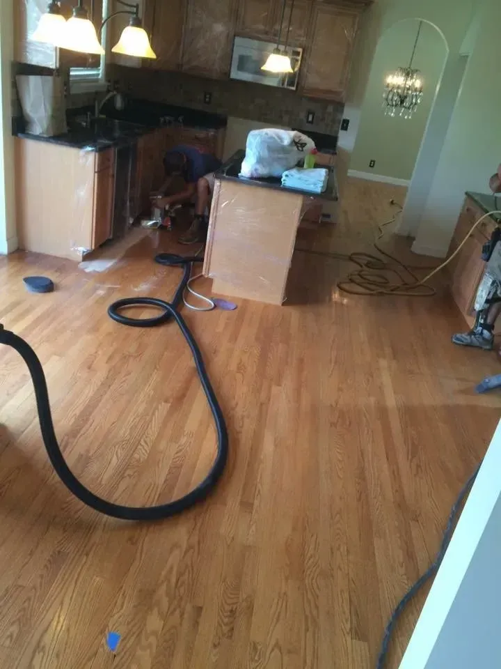 Hardwood floor installation in a kitchen with a worker, cabinets, and a vacuum hose.
