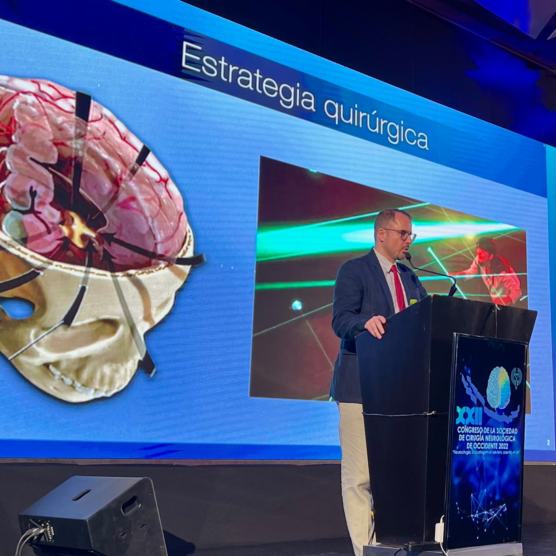 Un hombre se encuentra en un podio frente a una pantalla grande que dice estrategia quirúrgica