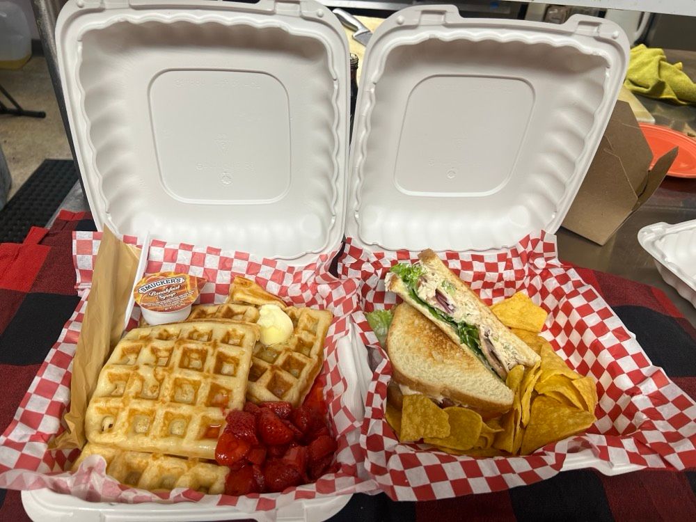 Two styrofoam containers filled with sandwiches , waffles , strawberries and chips.