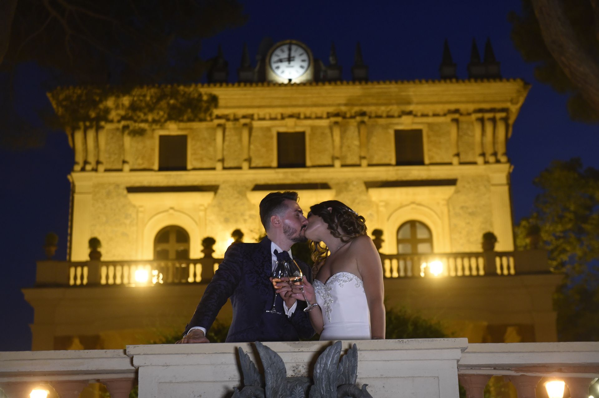 Una sposa e uno sposo si baciano davanti a un edificio con sopra un orologio.