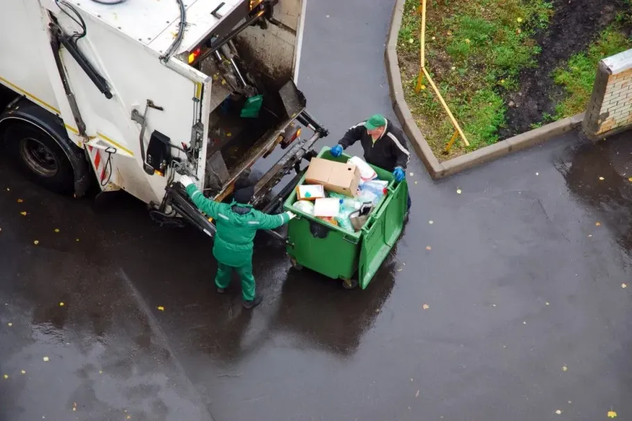 Worker in green uniform empties a trash bin into a garbage truck on a wet street.