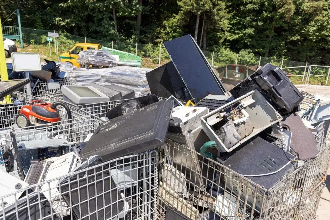 Pile of discarded electronics in wire bins outdoors, with broken black monitors and scrap metal.